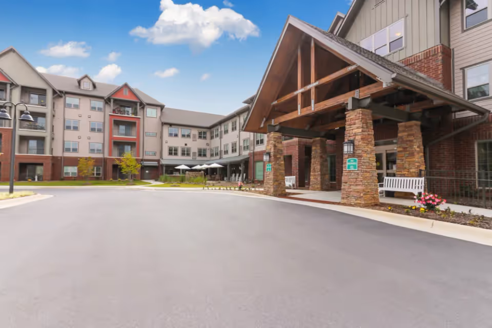 Exterior view of a senior living facility with a covered entrance supported by stone pillars, benches on the porch, and a multi-story building with balconies and windows under a blue sky with some clouds.