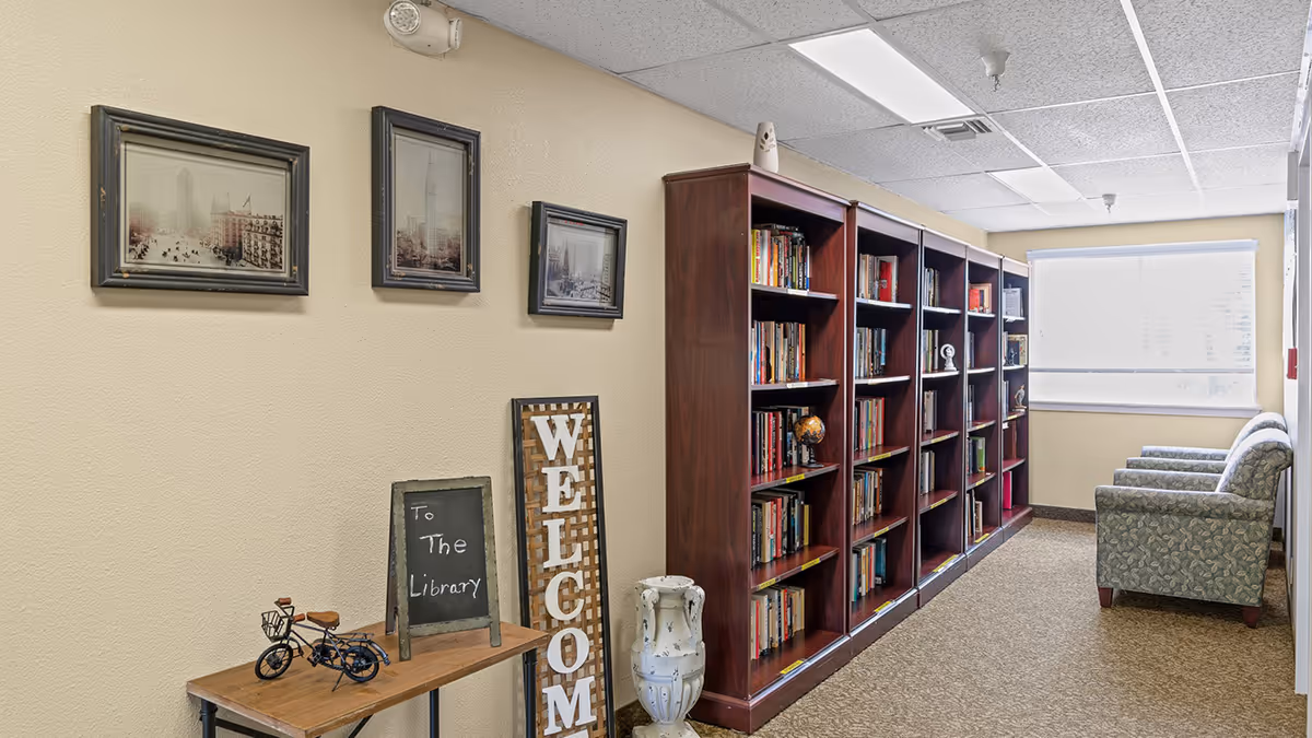 Interior hallway of Holiday Colonial Village featuring a wooden bookshelf filled with books, a small table with a miniature bicycle and a chalkboard sign that reads 'To The Library', a vertical 'WELCOME' sign, three framed pictures on the wall, and two upholstered armchairs near a window with a white blind.