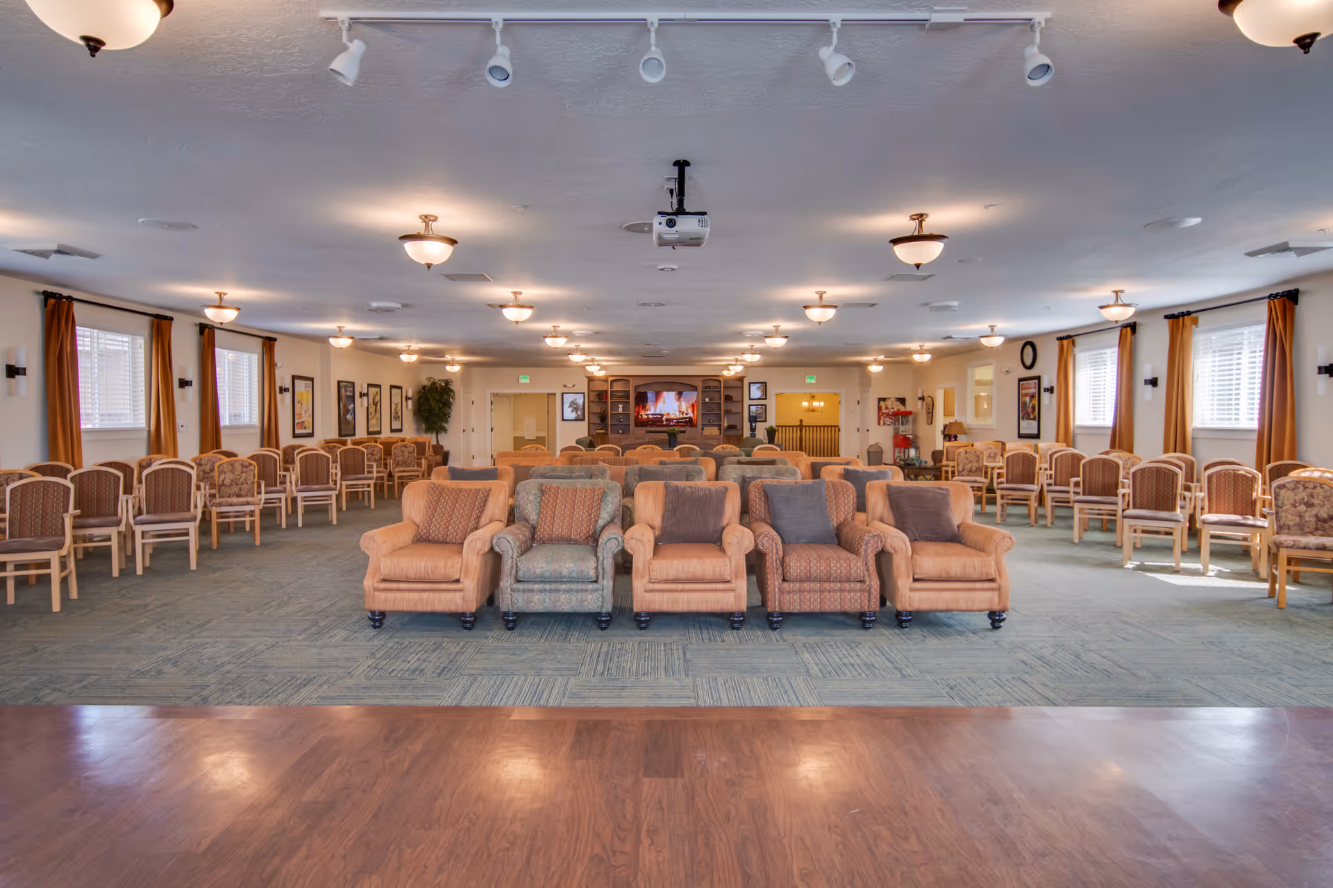 Large senior living community room with rows of chairs and a front row of upholstered armchairs facing a TV and ceiling-mounted projector.