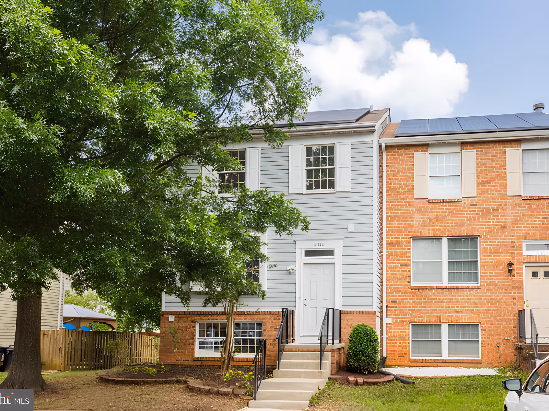 Exterior view of a two-story townhouse with light gray siding and white trim next to a red brick townhouse. There are stairs leading up to a white front door, a large green tree on the left side, and a small bush near the stairs. The sky is partly cloudy.