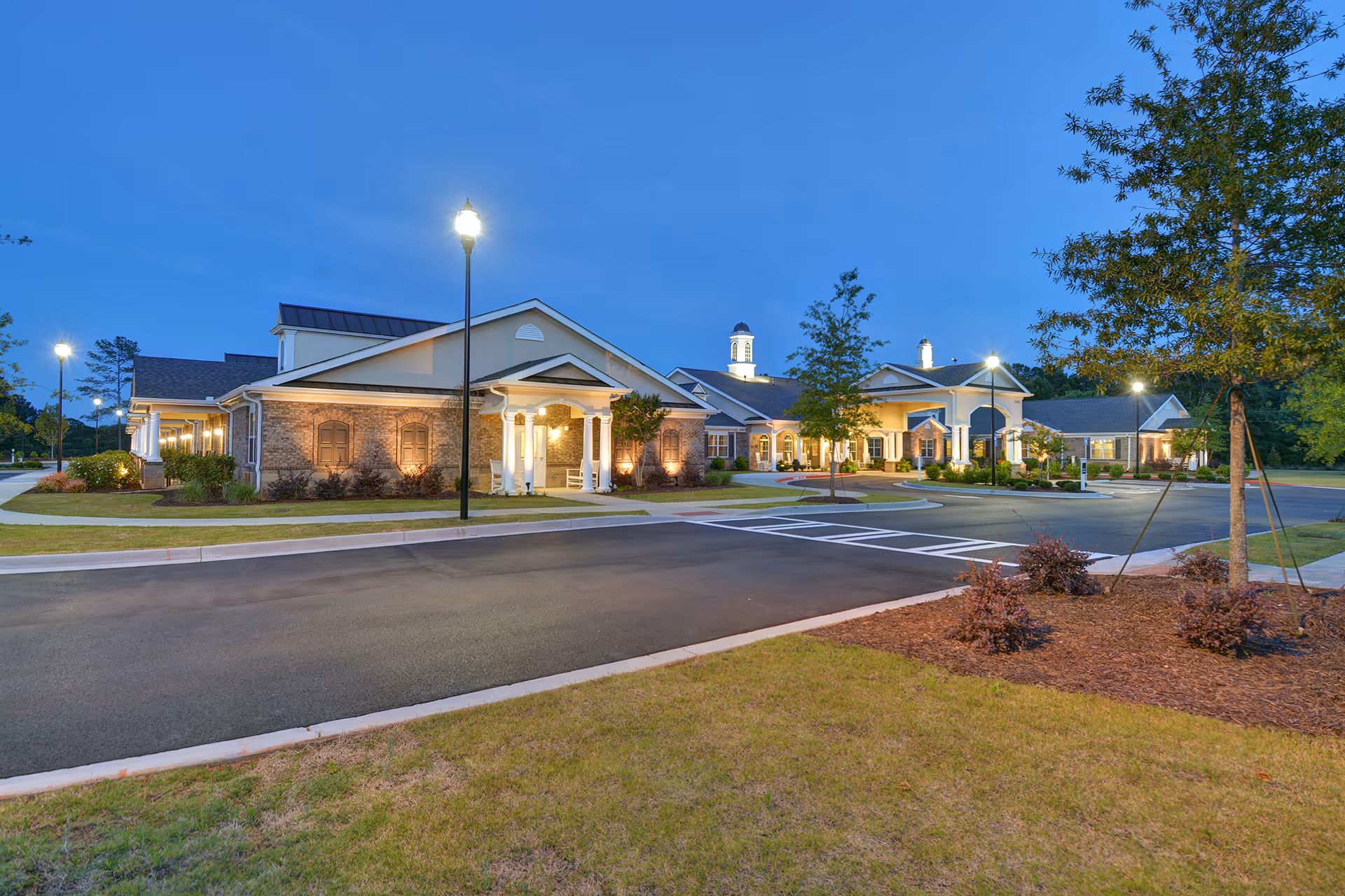 Exterior view of Benton Manor Of Stockbridge at dusk, showing a well-lit building with brick and siding facade, multiple entrances, street lamps, landscaped grounds, and a paved driveway.