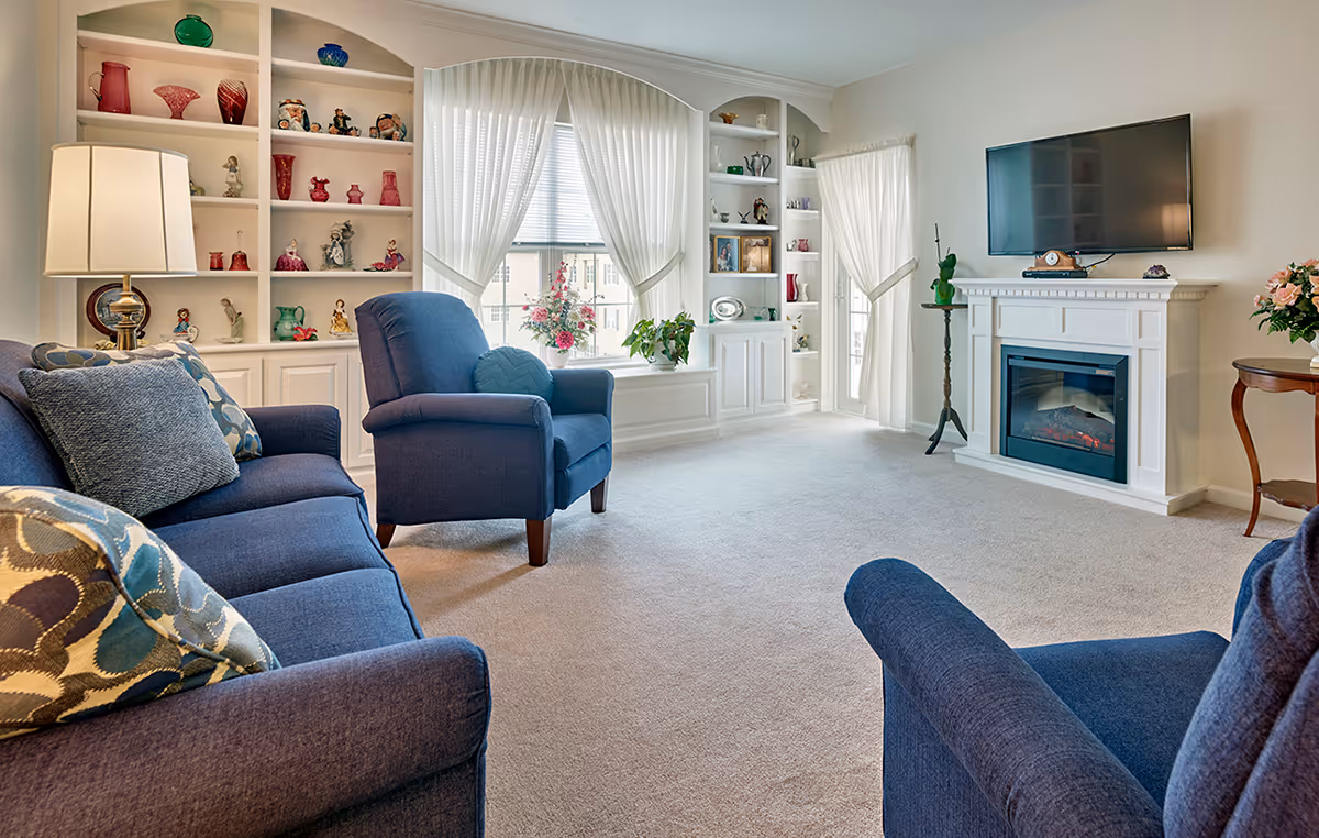 A bright and cozy living room with blue upholstered sofas and armchairs, a white fireplace with a mounted flat-screen TV above it, built-in white shelves filled with decorative items, and large windows with sheer white curtains letting in natural light.