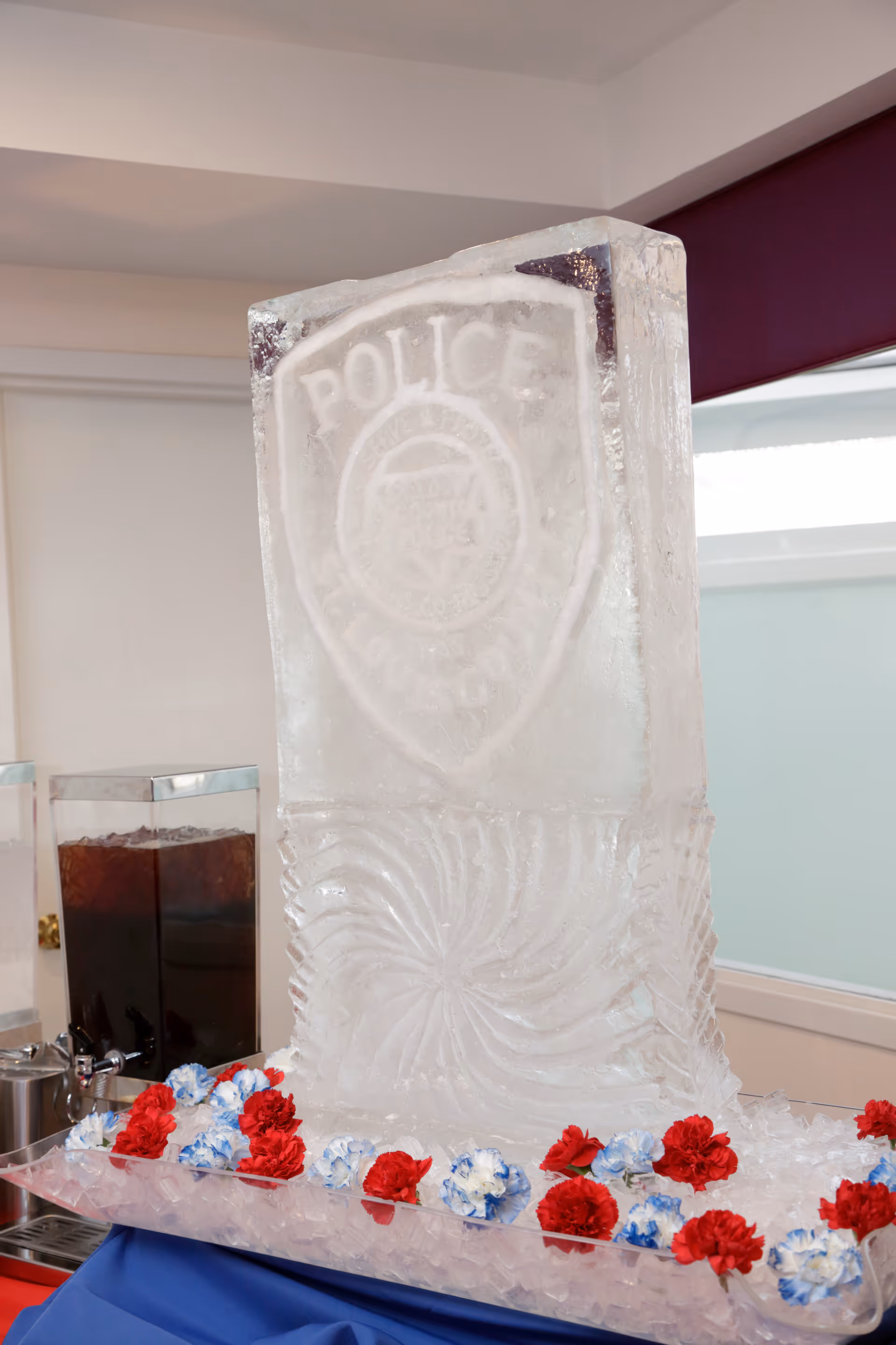 An ice sculpture featuring a police badge emblem, displayed on a bed of ice with red, white, and blue flowers around the base. The sculpture is indoors, with a beverage dispenser visible in the background.