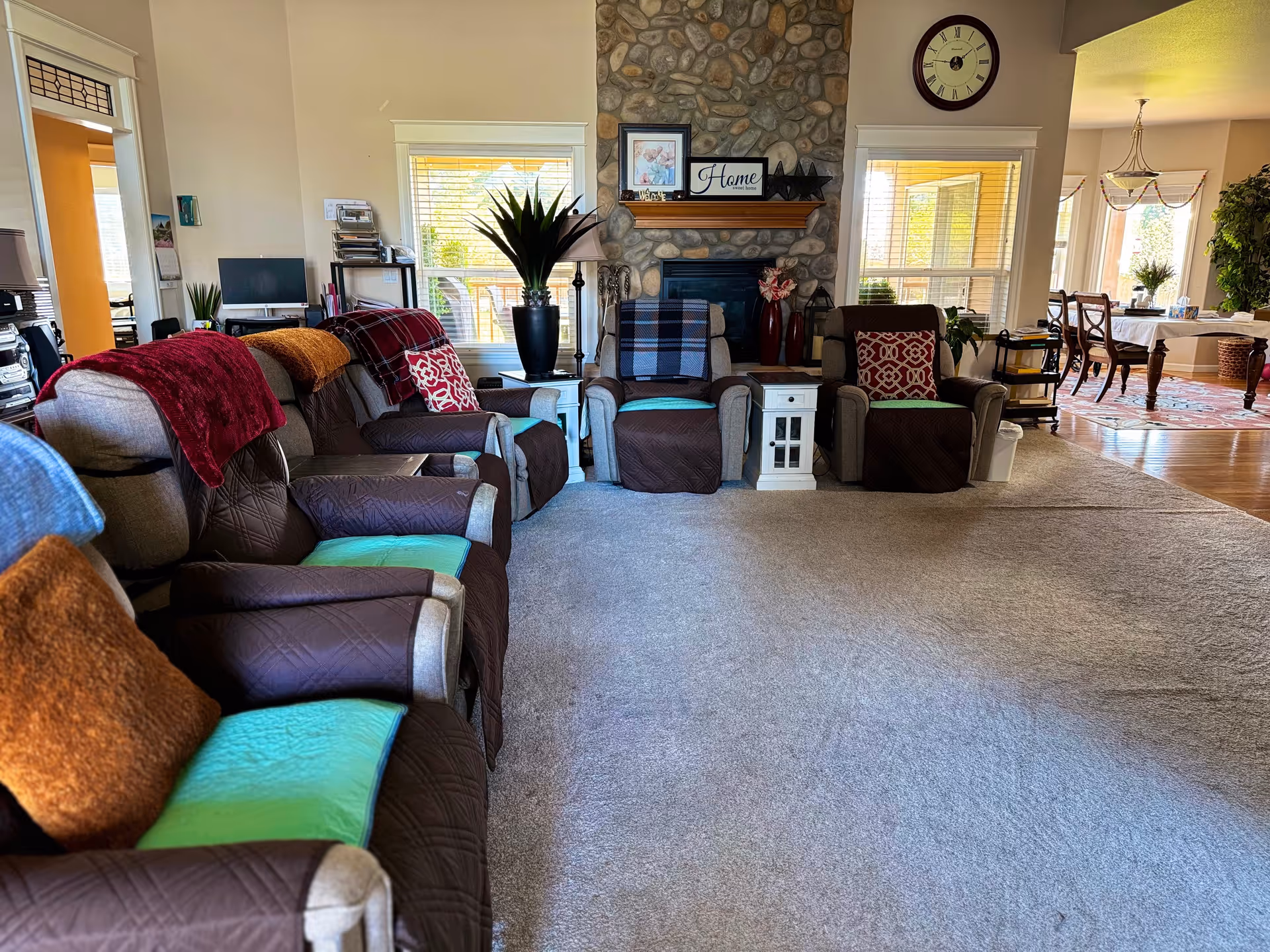 A cozy living room with several cushioned armchairs arranged in a semi-circle facing a stone fireplace. The chairs have colorful blankets and pillows on them. There are two windows on either side of the fireplace letting in natural light. A clock hangs above the fireplace mantel, which holds decorative items including a framed picture and a sign that says 'Home'. To the right, a dining area with a table and chairs is visible, along with a chandelier and a large plant.