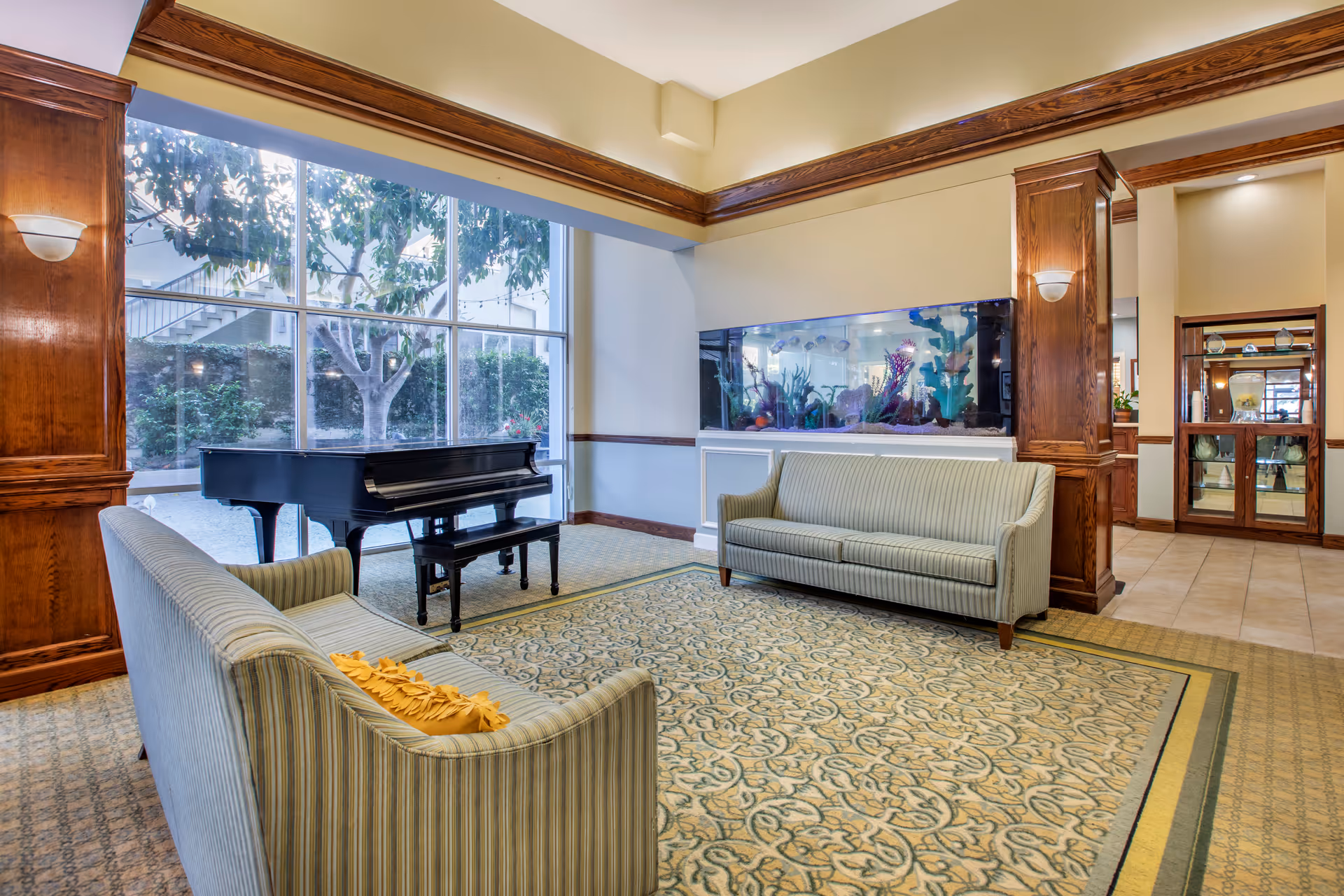 A cozy living room area with two striped sofas facing each other on a patterned carpet. A black grand piano is positioned near large windows showing outdoor greenery. There is a large built-in aquarium with colorful coral and fish on one wall, and wooden pillars and trim add warmth to the space.