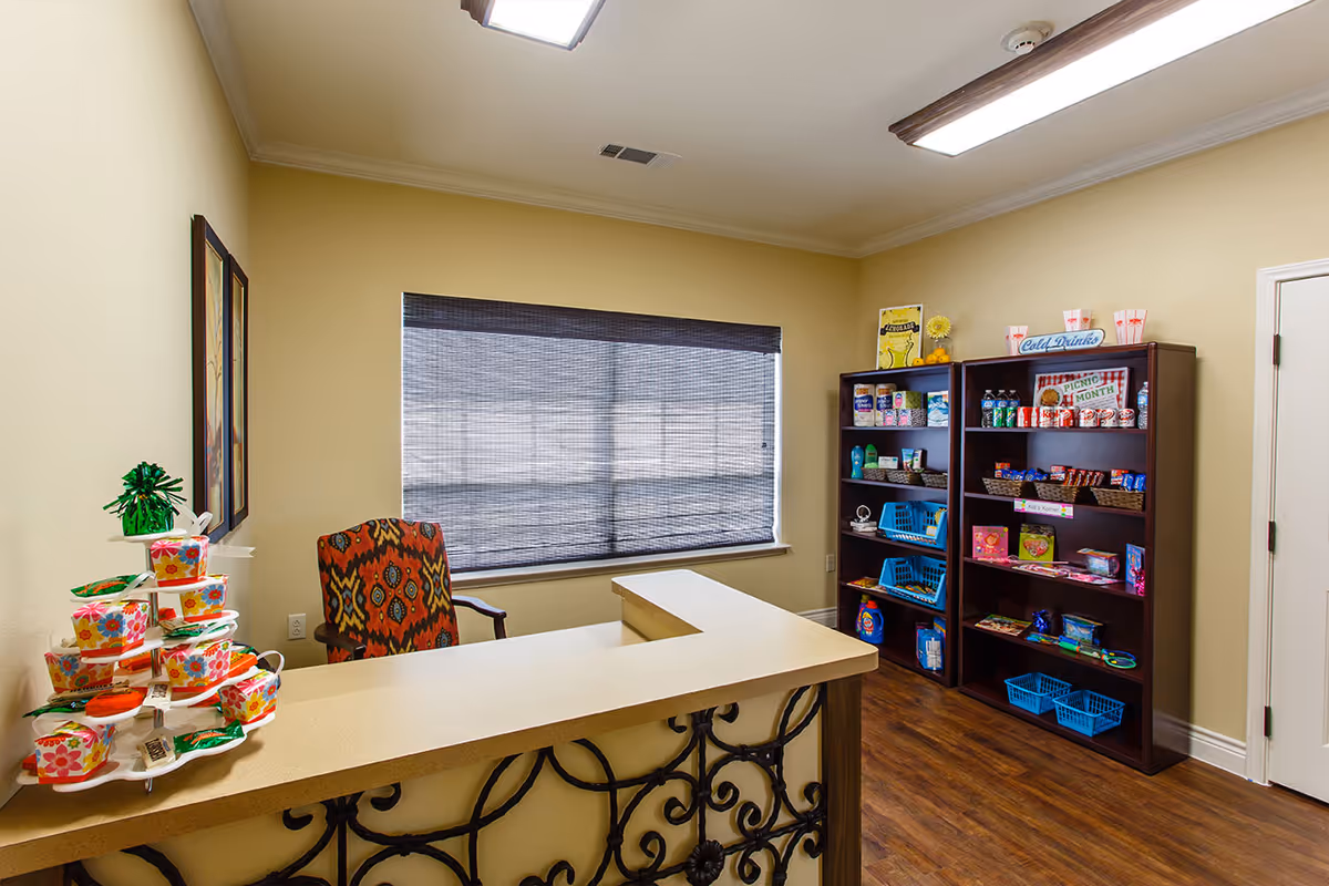 Interior view of a small snack and refreshment area in a senior living facility. There is a counter with a decorative iron front and a colorful patterned chair behind it. On the counter, there is a tiered display with small colorful boxes and packets. Against the wall, there are two dark wooden shelves stocked with various snacks, drinks, and personal care items. A window with a dark woven shade is centered on the back wall, and the room has warm beige walls and wood flooring.