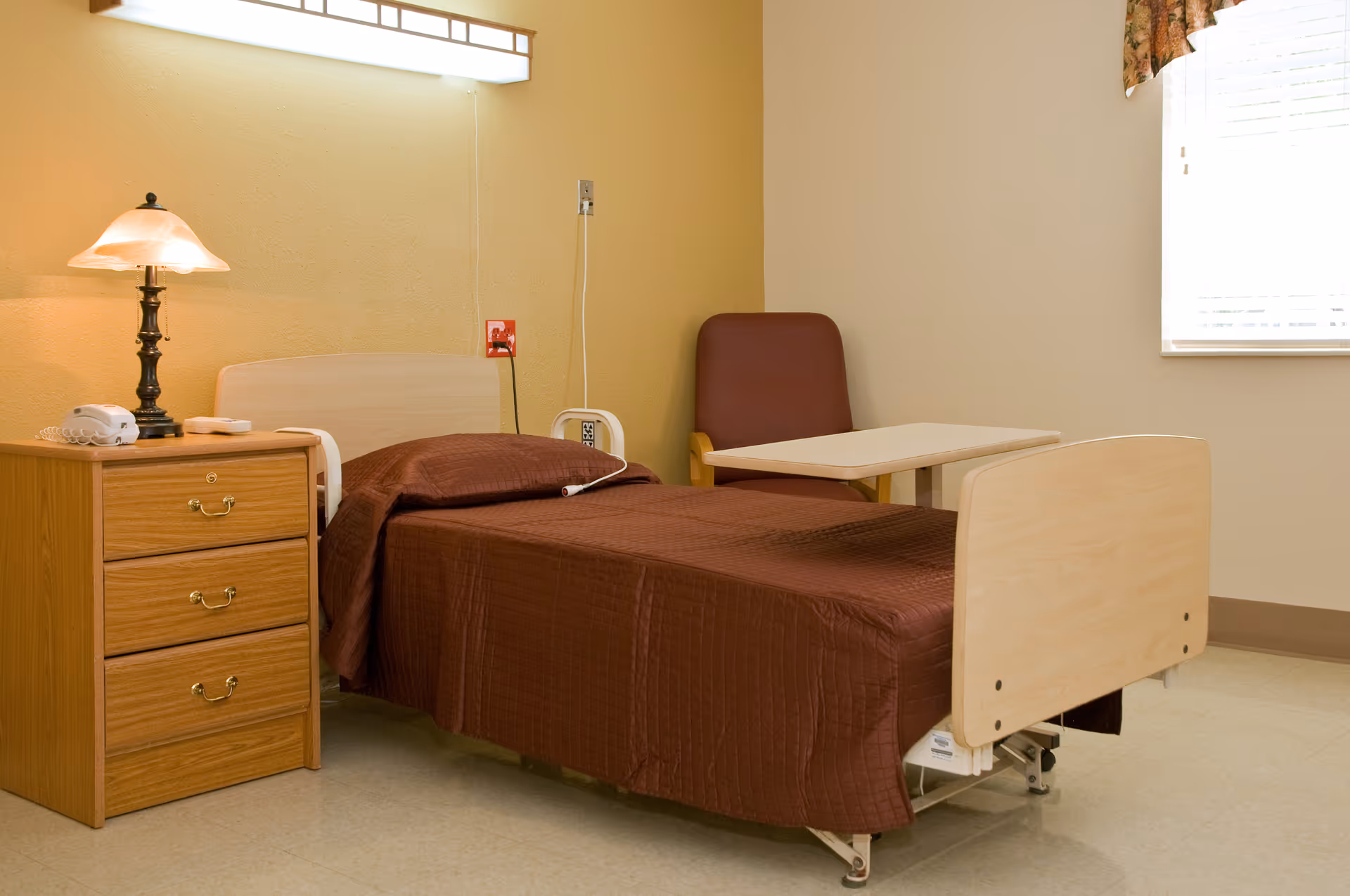 A simple senior living facility bedroom with a single bed covered in a brown quilt, a wooden nightstand with three drawers, a lamp, and a telephone. There is a maroon chair and an overbed table next to the bed, and a window with blinds and a floral valance letting in natural light.