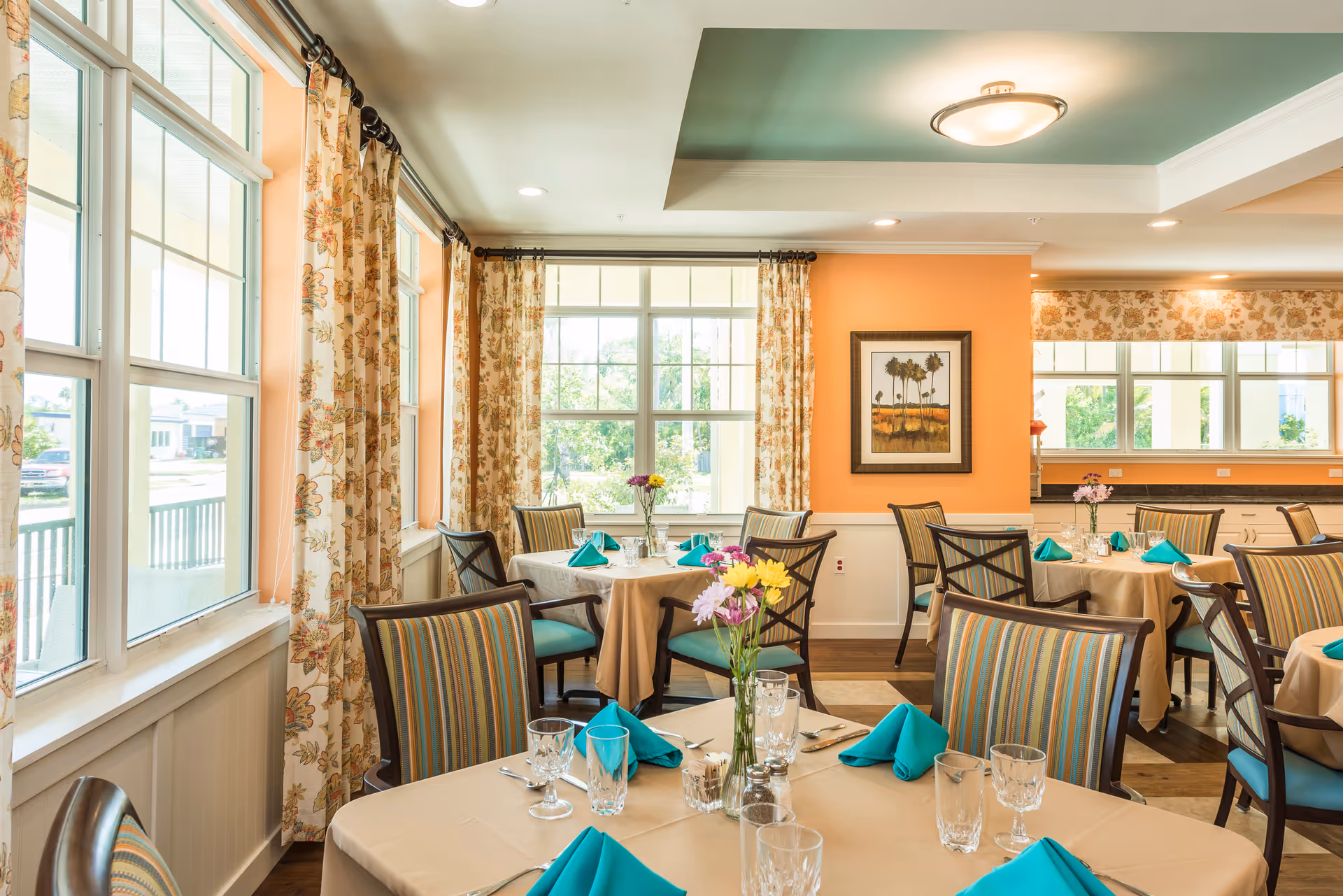 Bright dining room with multiple tables covered in beige tablecloths, each set with turquoise napkins, glassware, and silverware. The room features large windows with floral curtains, striped cushioned chairs, and a framed painting on a peach-colored wall. Fresh flowers in vases are placed on the tables.