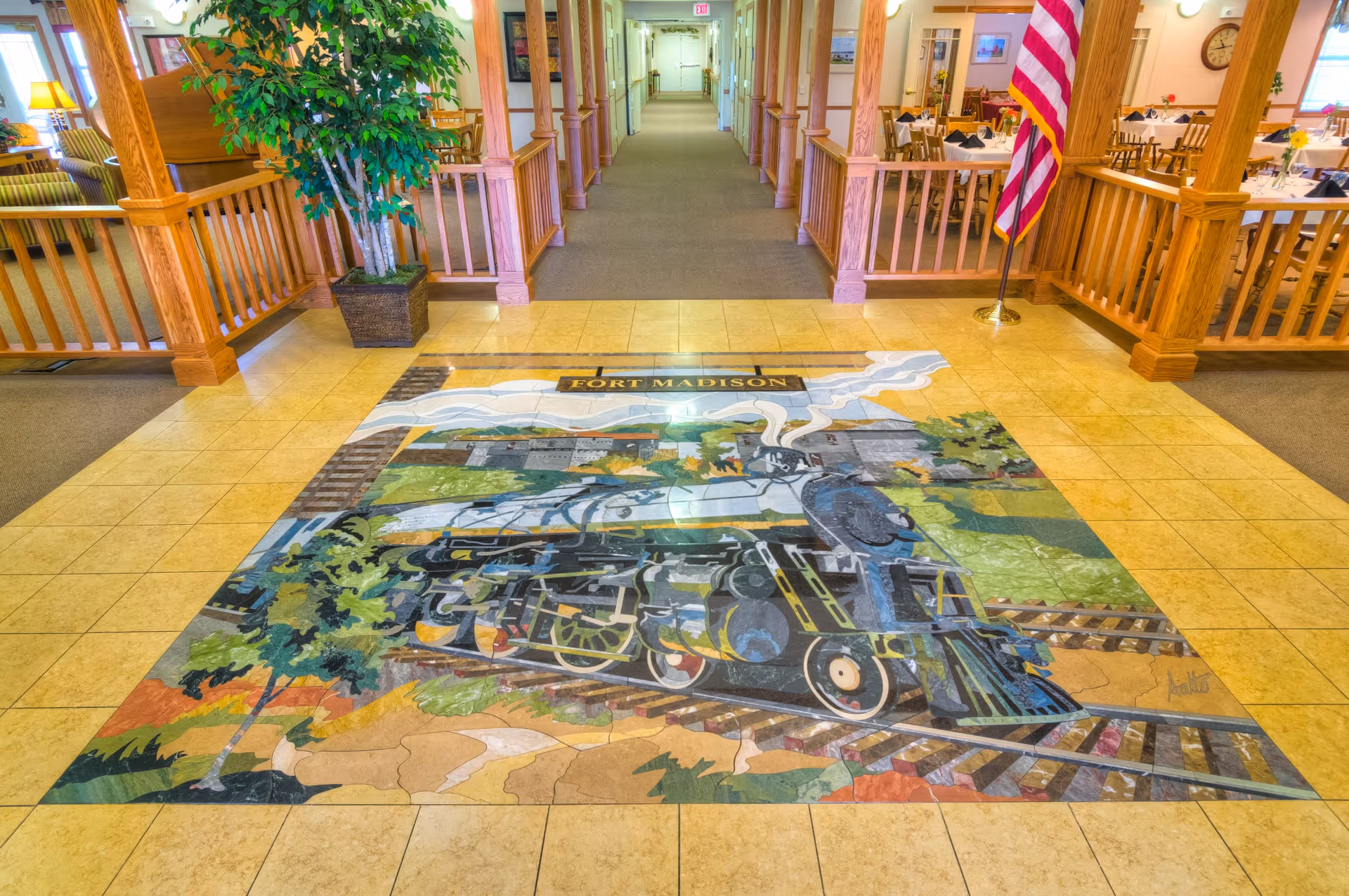 Interior hallway of a senior living facility with wooden railings and columns on both sides. The floor features a large colorful mosaic artwork of a steam locomotive train with the text 'Fort Madison'. There is a potted plant on the left and an American flag on the right near the entrance to a dining area with tables and chairs.