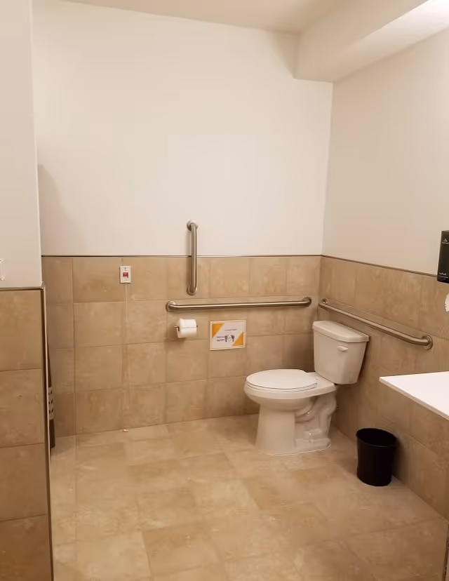 Accessible restroom with a toilet, stainless steel grab bars, tiled walls and floor, and part of a wall-mounted sink visible.