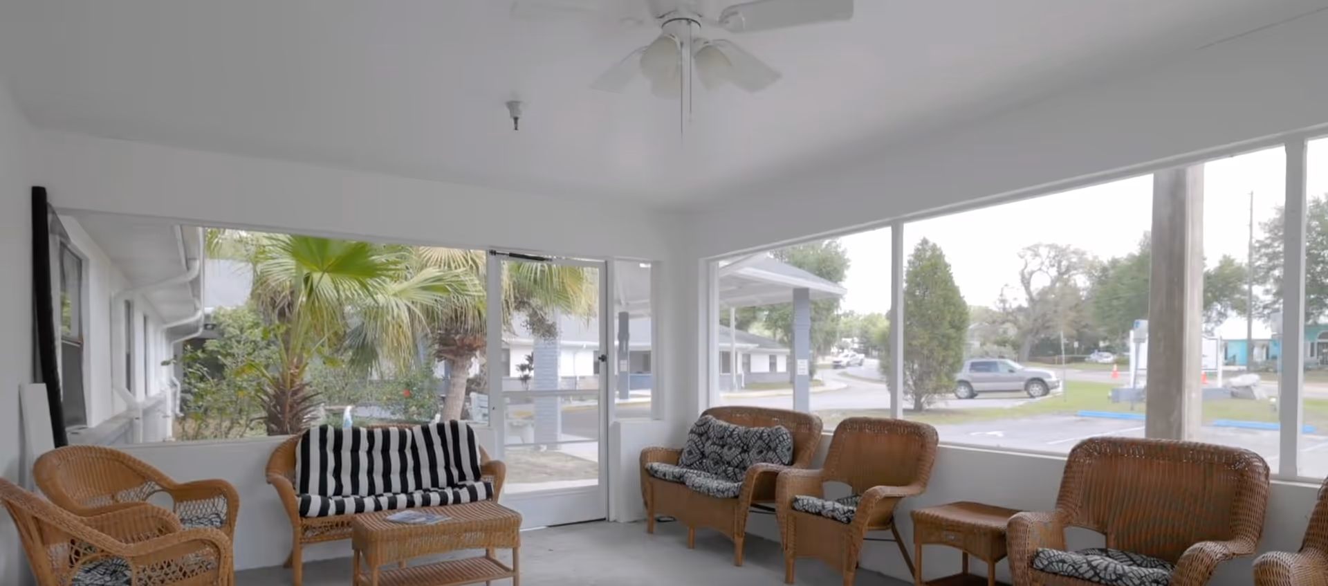 Bright enclosed sunroom with wicker chairs and a loveseat arranged around a small table and large windows looking out to palm trees and a street.