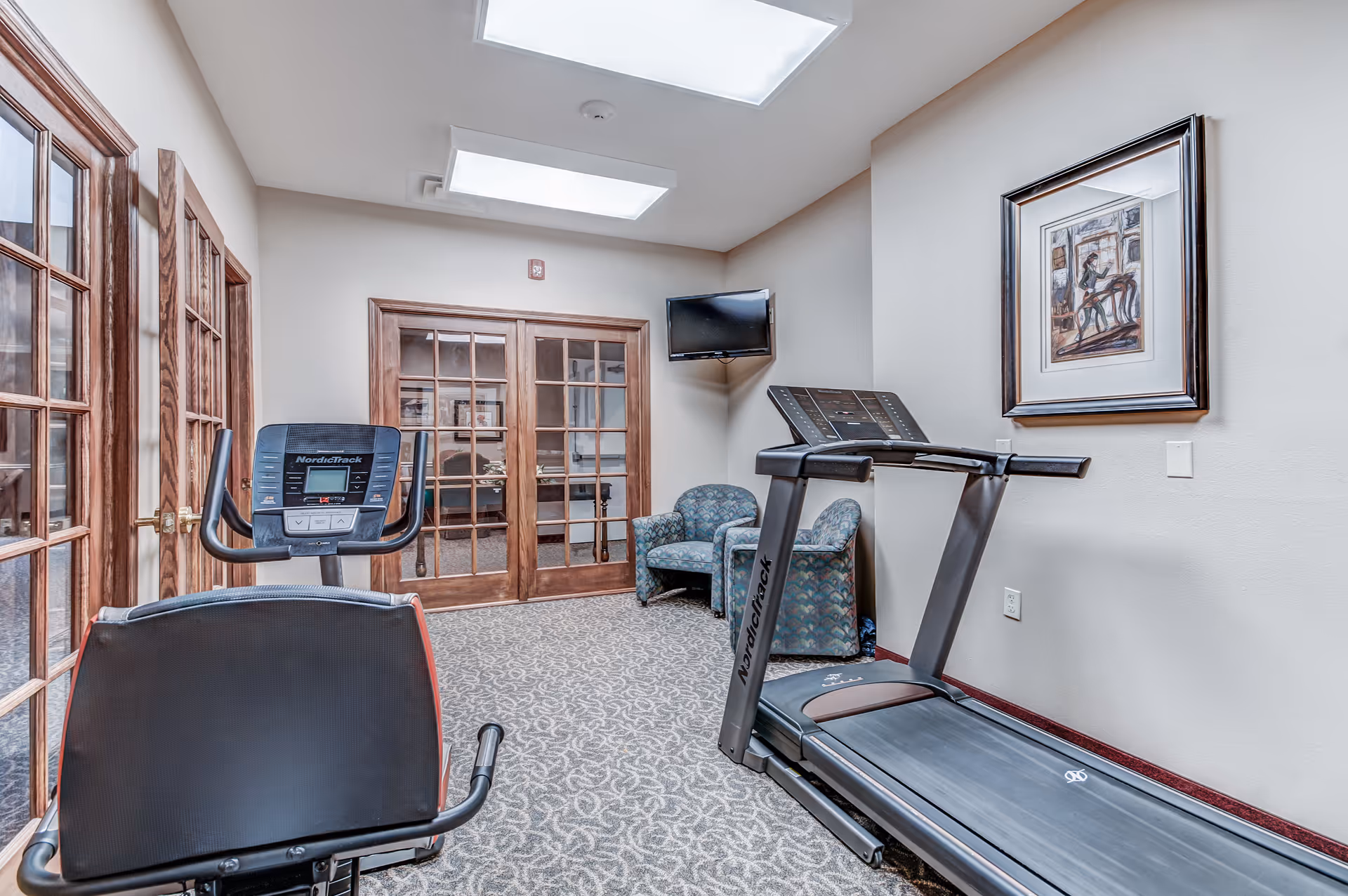 A small fitness room with a treadmill and an exercise bike facing a set of wooden-framed glass double doors. There are two patterned armchairs in the corner beneath a wall-mounted TV and a framed artwork on the wall. The room has carpeted flooring and bright overhead lighting.