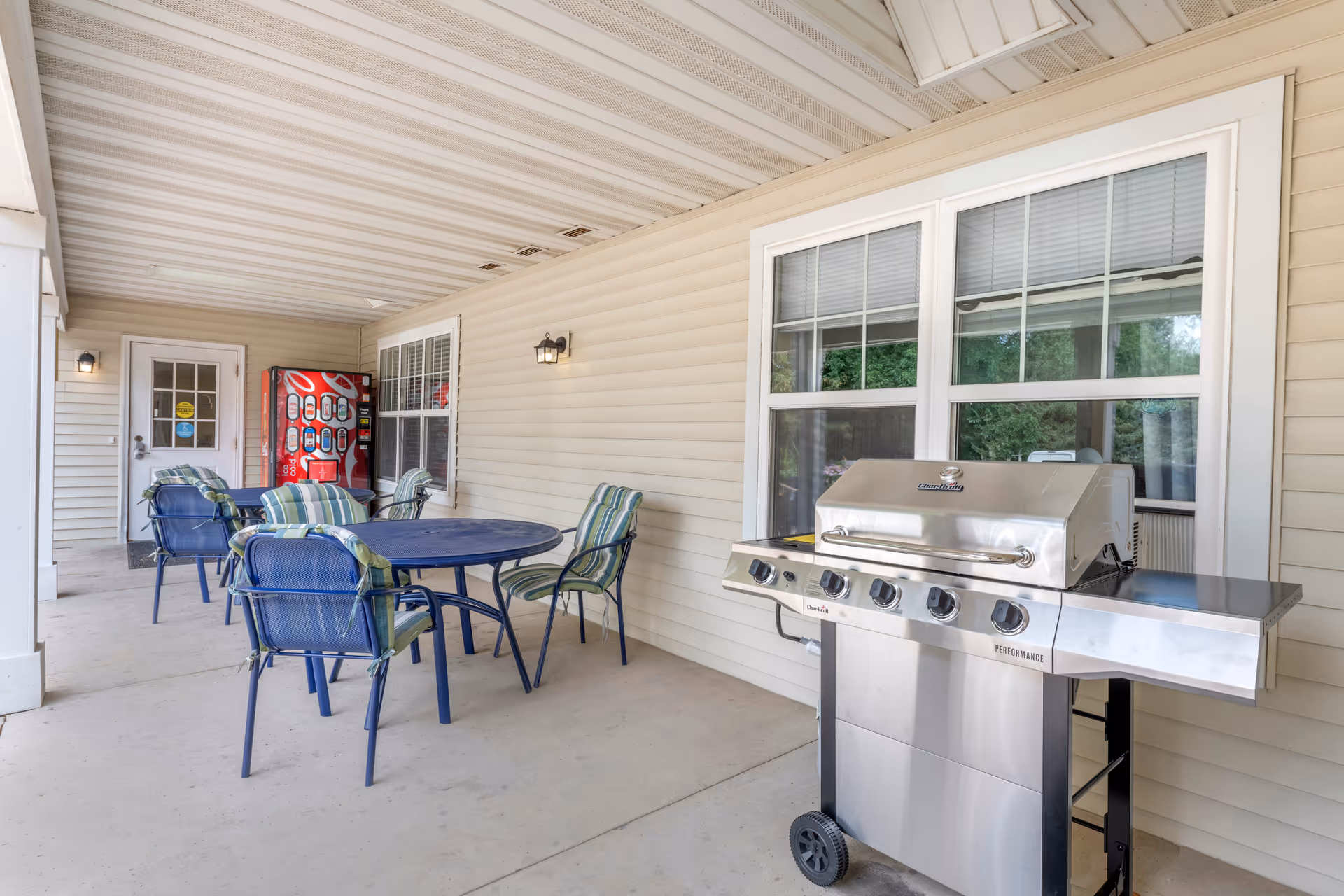 Covered outdoor patio area with a stainless steel gas grill on the right, a round blue metal table with four blue metal chairs with green and white striped cushions, and a red vending machine near a white door in the background.