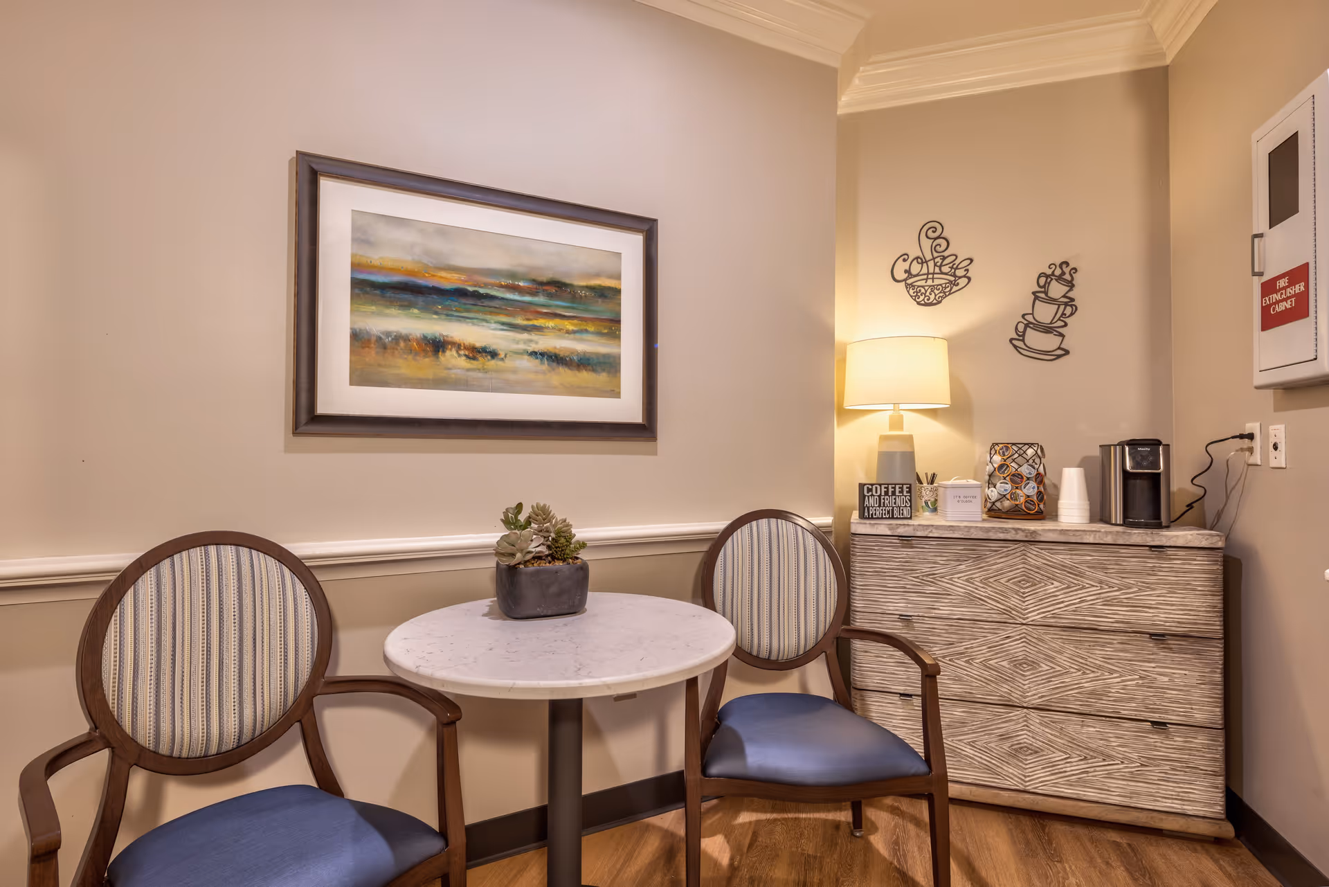 A small seating area with two wooden chairs with blue cushions and striped backs around a round white table with a small plant centerpiece. On the wall above the table is a framed abstract landscape painting. To the right, there is a wooden cabinet with a lamp, coffee pods, a coffee maker, and decorative wall art shaped like coffee cups. A fire extinguisher cabinet is mounted on the wall above the cabinet.