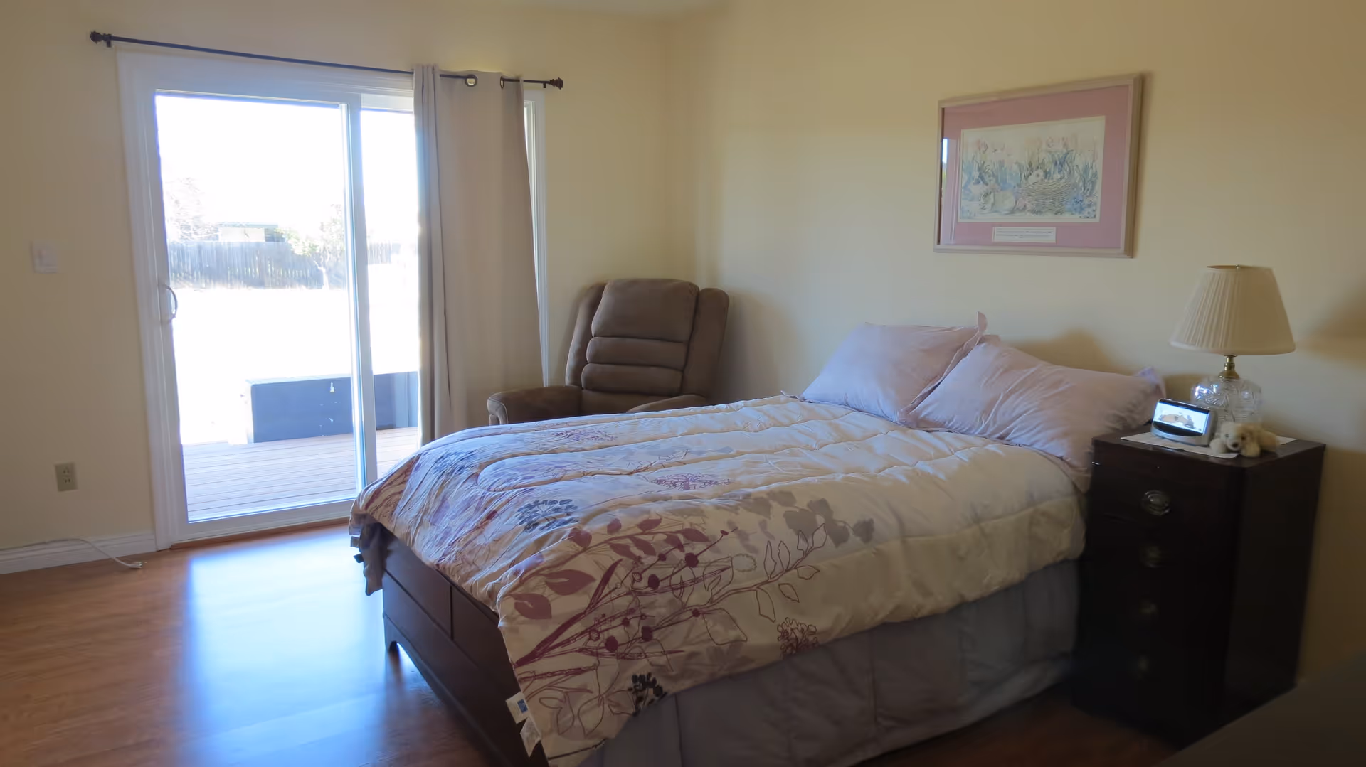 A bedroom with a bed covered in a floral-patterned comforter, two pillows, a brown recliner chair in the corner, a wooden nightstand with a lamp, a small digital clock, and a stuffed animal. There is a framed picture on the wall and a sliding glass door with curtains leading outside.
