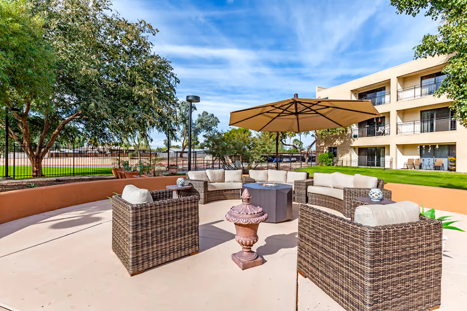 Outdoor patio area with wicker furniture including cushioned chairs and sofas arranged around a central fire pit table under a large beige umbrella. The patio is surrounded by trees, a fence, and a multi-story building with balconies in the background under a partly cloudy blue sky.