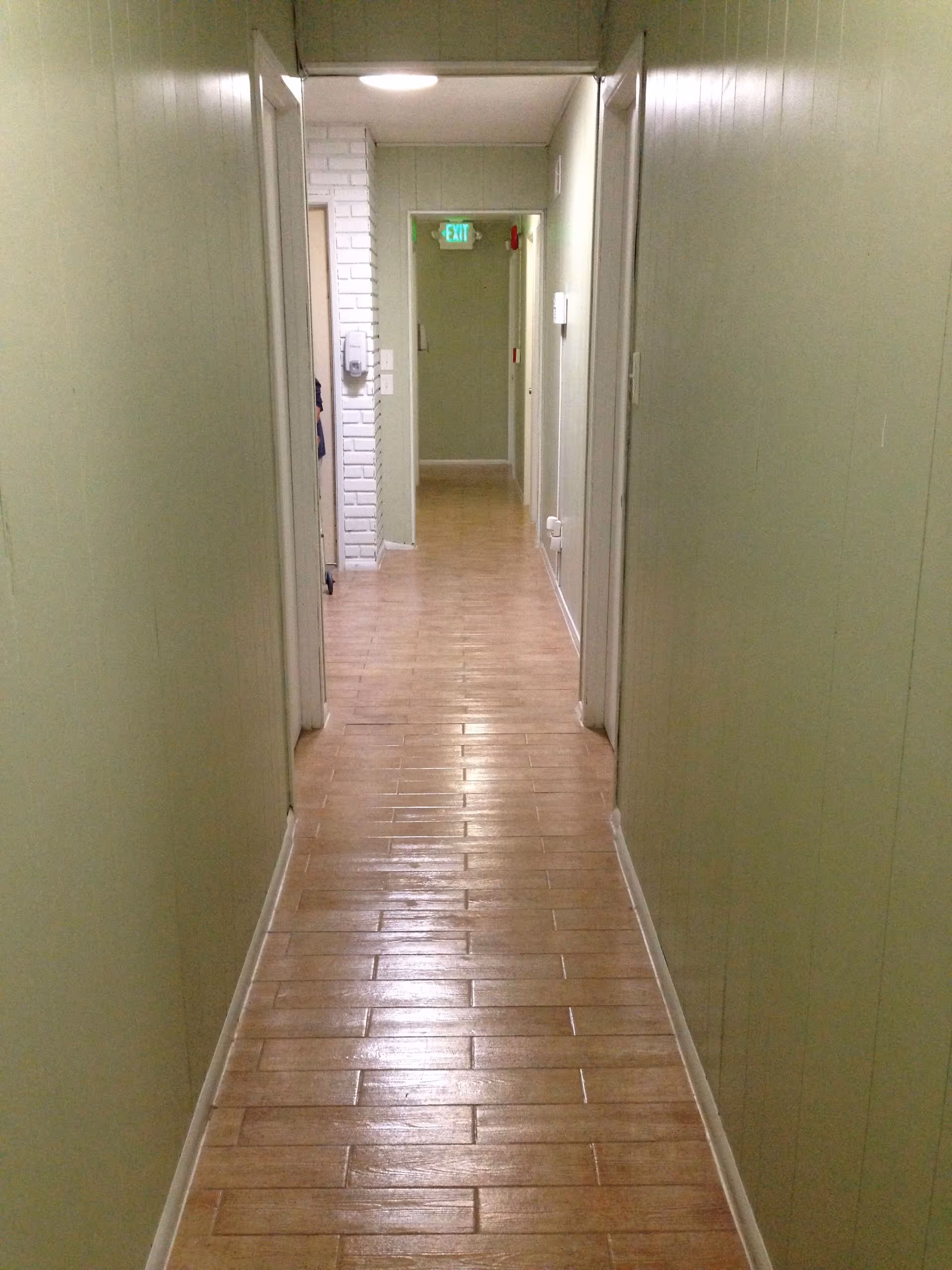 A narrow indoor hallway with light green walls and a wooden tile floor. There are several doorways along the hallway, and an exit sign is visible at the far end above a door.