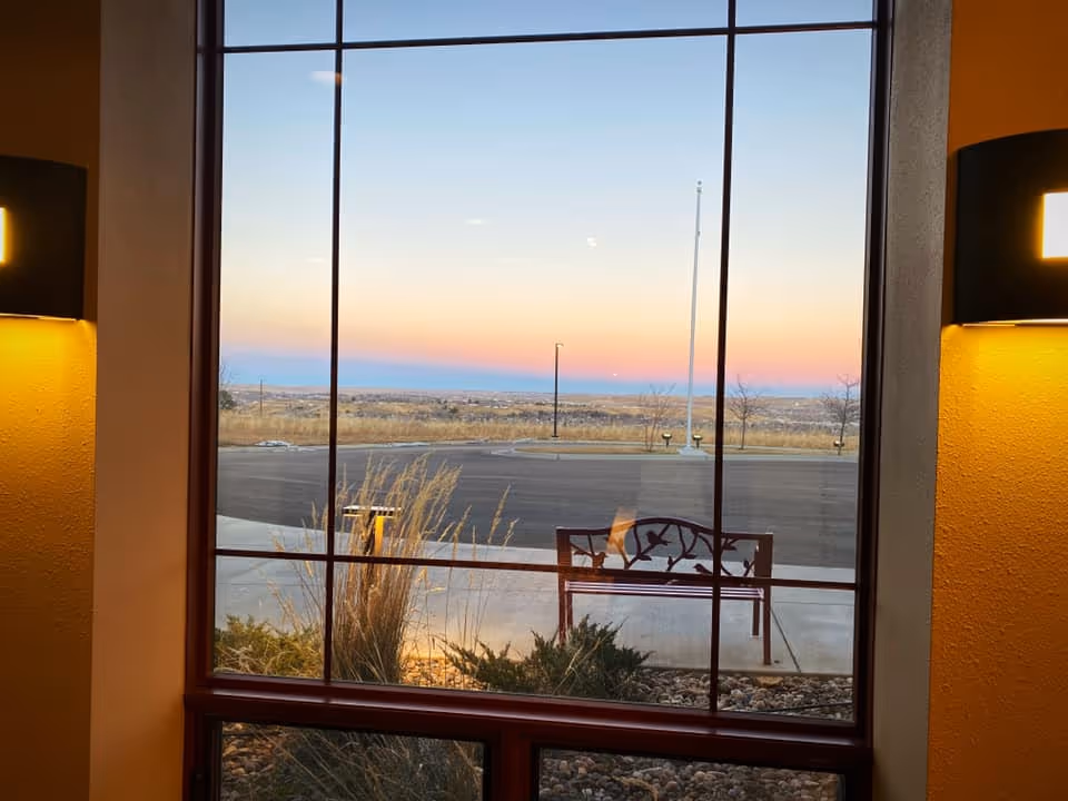 View through a large window showing an outdoor bench with a decorative backrest, some plants, a parking lot, and a sunset sky with gradient colors from blue to pink.