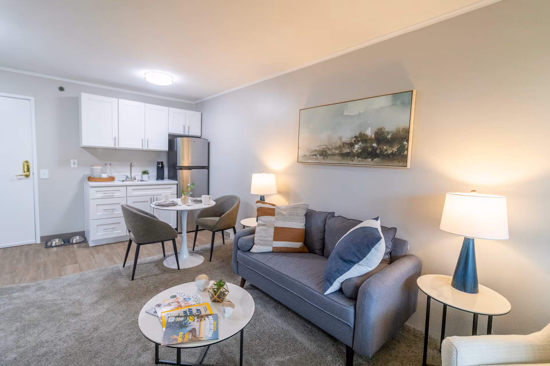 A cozy living area with a gray sofa adorned with three pillows, two side tables each with a lamp, a round coffee table with magazines and decorative items, and a small dining area with a round table and two chairs. In the background, there is a compact kitchen with white cabinets, a stainless steel refrigerator, a sink, and a coffee maker. The walls are light-colored, and a framed landscape painting hangs above the sofa.
