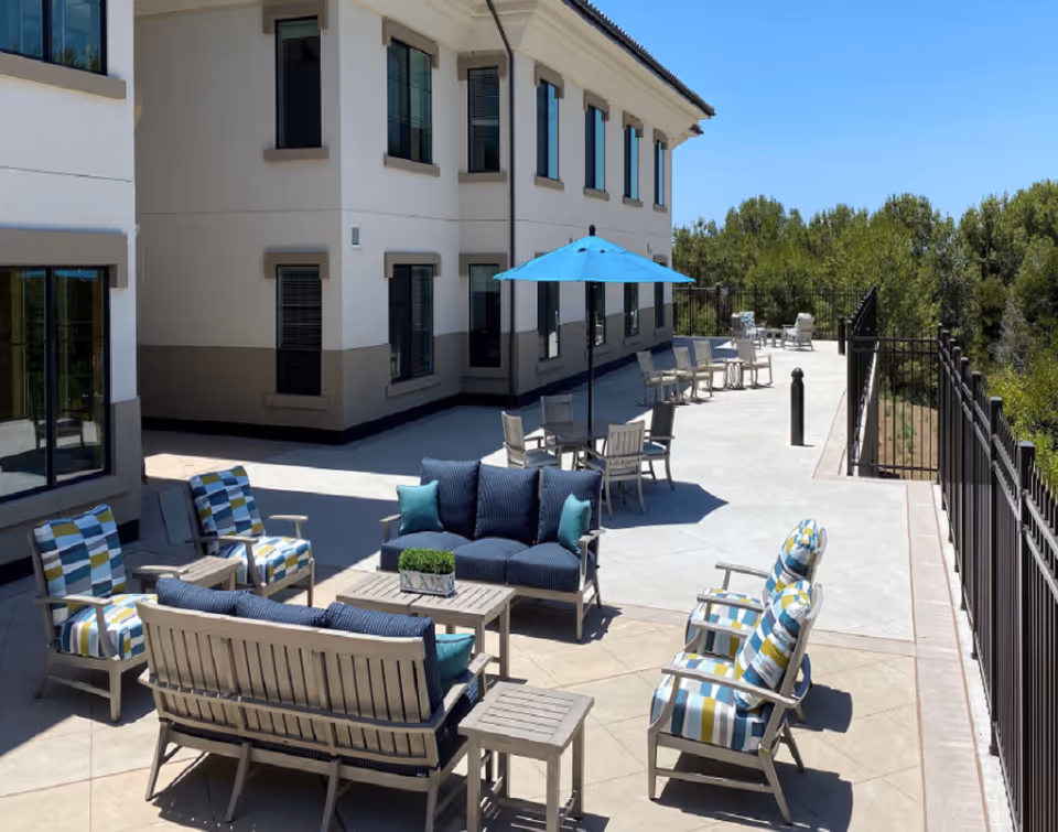 Outdoor patio area at Silvergate Rancho Bernardo featuring cushioned wooden chairs and sofas with striped and solid blue pillows, a table with a small plant, and a blue umbrella providing shade over a dining table. The patio is adjacent to a two-story building with multiple windows and is surrounded by a black metal fence with trees in the background under a clear blue sky.