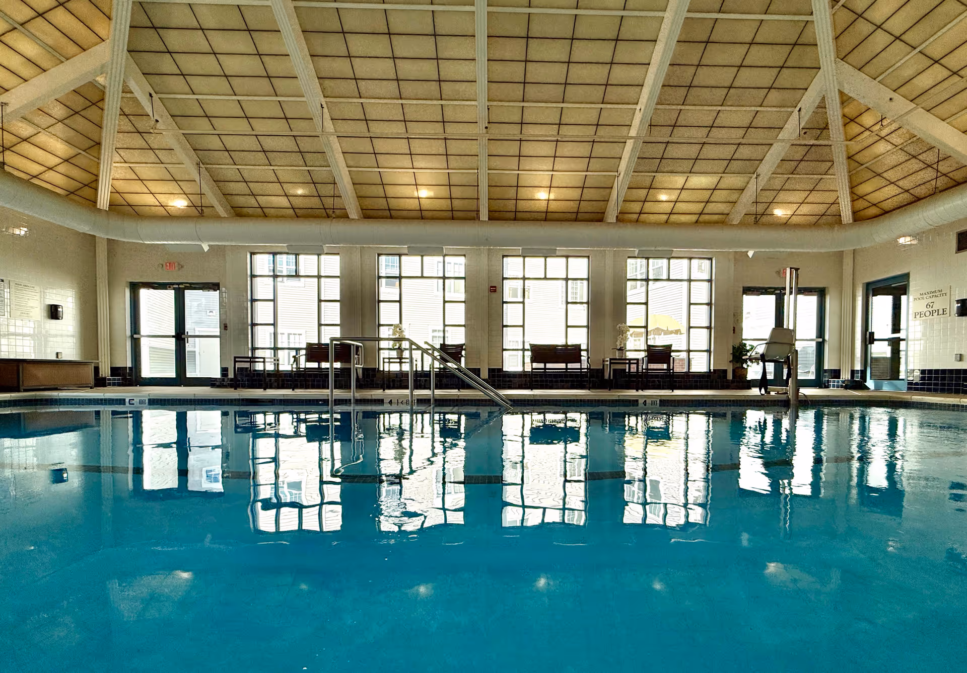 Indoor swimming pool with clear blue water, metal handrails, and pool lift chair. The room has large windows letting in natural light, a high ceiling with exposed beams, and seating areas with chairs and tables along the far wall.