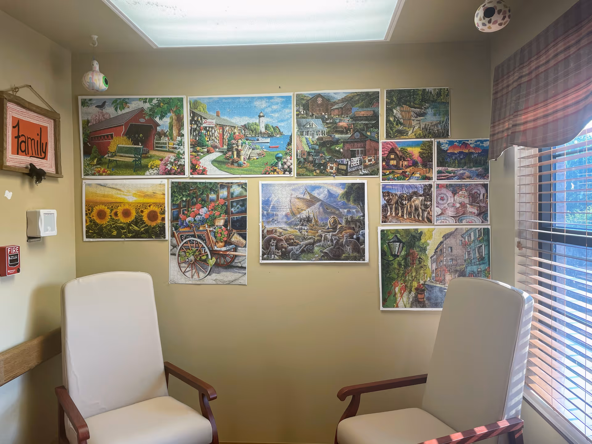 A cozy room with two white cushioned chairs with wooden armrests facing each other. The beige walls are decorated with multiple colorful paintings and pictures, including scenes of nature, animals, and landscapes. A window with blinds and a plaid valance allows natural light to enter the room. A small framed sign on the wall reads 'family'.