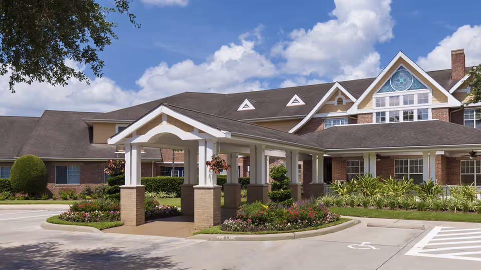 Covered porte-cochere entrance to a brick senior living building with landscaped flower beds under a blue sky.