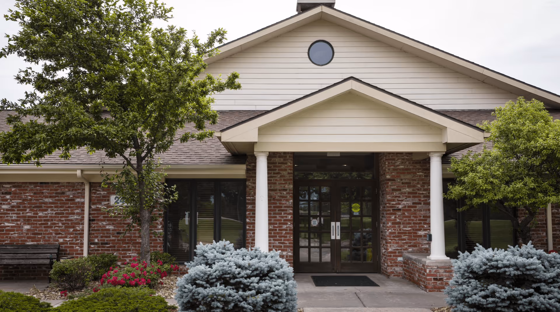 Front exterior view of a single-story brick building with a peaked roof, white columns flanking the entrance, glass double doors, and landscaping including trees, bushes, and flowers.