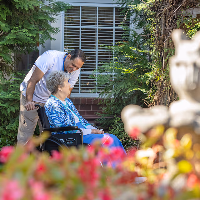A man in a white shirt is smiling and leaning over to talk to an elderly woman sitting in a wheelchair in a garden area with greenery and flowers around them.