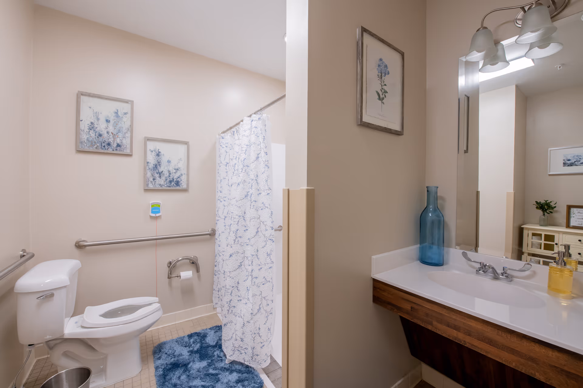 A clean and accessible bathroom with a white toilet, grab bars, a shower with a white and blue patterned curtain, a blue bath mat, and two framed floral pictures on the wall. The bathroom also features a vanity with a sink, a large mirror, a blue decorative vase, and a soap dispenser.