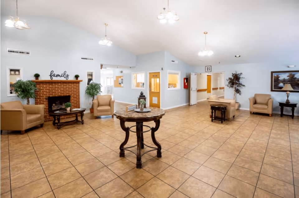 A spacious and well-lit common area in a senior living facility with beige armchairs arranged around a brick fireplace with the word 'family' above it. There is a round wooden table in the center with decorative items, tile flooring, light blue walls, and ceiling lights. The room has multiple doorways and windows, with plants and framed artwork adding to the decor.