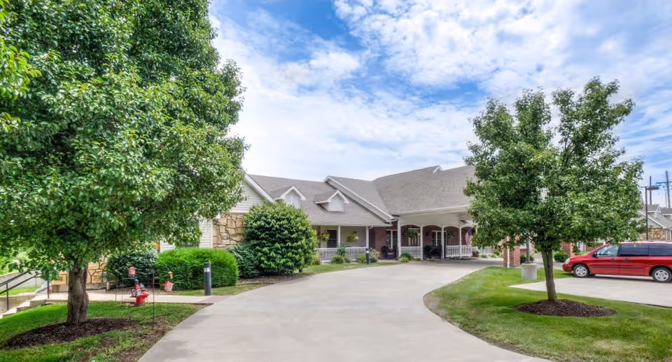 Exterior view of a senior living facility with a curved driveway leading to the entrance. The building has a covered porch with white railings and brick columns. There are green trees and shrubs on either side of the driveway, and a red minivan is parked on the right side. The sky is partly cloudy.