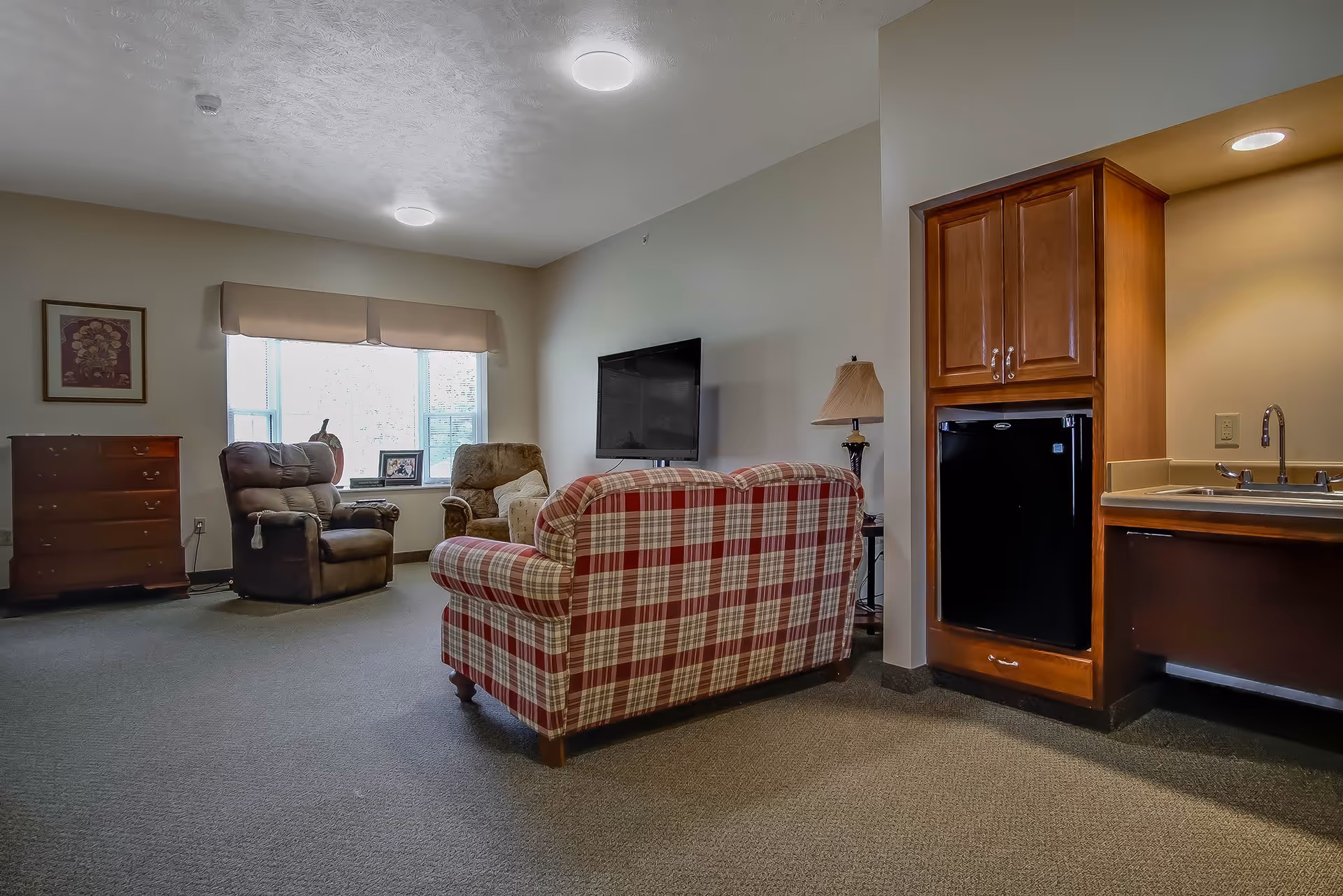 A cozy living area in a senior living facility featuring a plaid sofa facing a wall-mounted flat screen TV. There are two armchairs near a window with beige valance curtains. A wooden chest of drawers and a framed picture are on the left wall. On the right side, there is a small kitchenette area with wooden cabinets, a mini fridge, a sink, and a lamp on a side table next to the sofa. The room has neutral-colored walls and carpeted flooring.