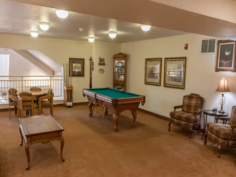 A cozy common area with a pool table in the center, two plaid upholstered chairs with a small round table and lamp between them, a wooden coffee table, and a card table with four chairs near a railing overlooking a lower level. The walls are decorated with framed pictures and a wooden grandfather clock.