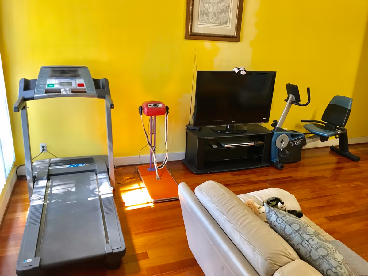 A room with yellow walls and wooden flooring containing exercise equipment including a treadmill and a stationary bike, a black TV stand with a flat-screen TV, and a beige sofa with patterned cushions and a stuffed animal.