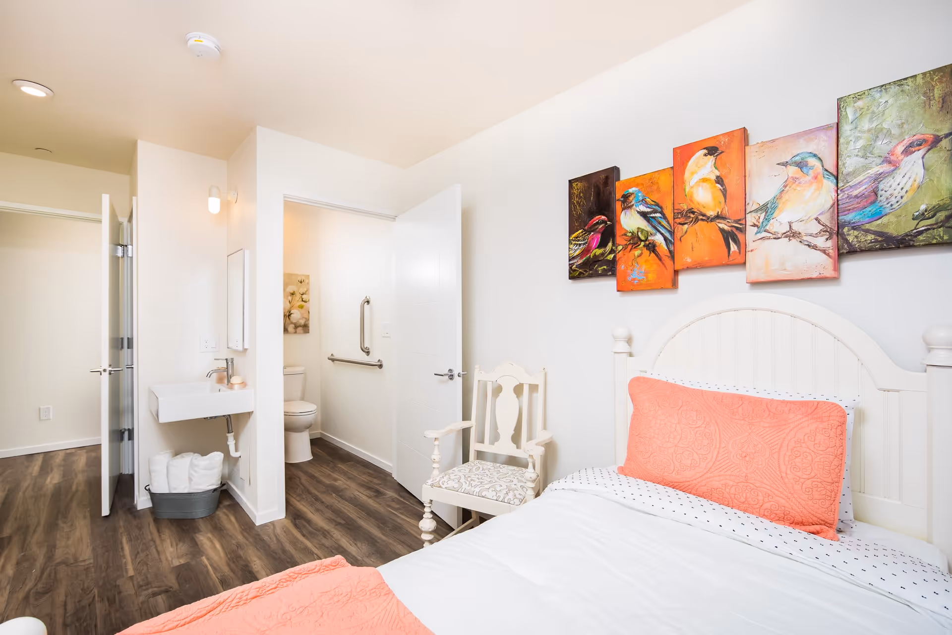 A bright bedroom with a white bed featuring a coral-colored pillow and blanket. Above the bed, there are five colorful paintings of birds. A white wooden chair with a patterned cushion is placed next to the bed. The room has wood flooring and white walls. An open door reveals a bathroom with a toilet, grab bars, and a small sink with towels underneath.