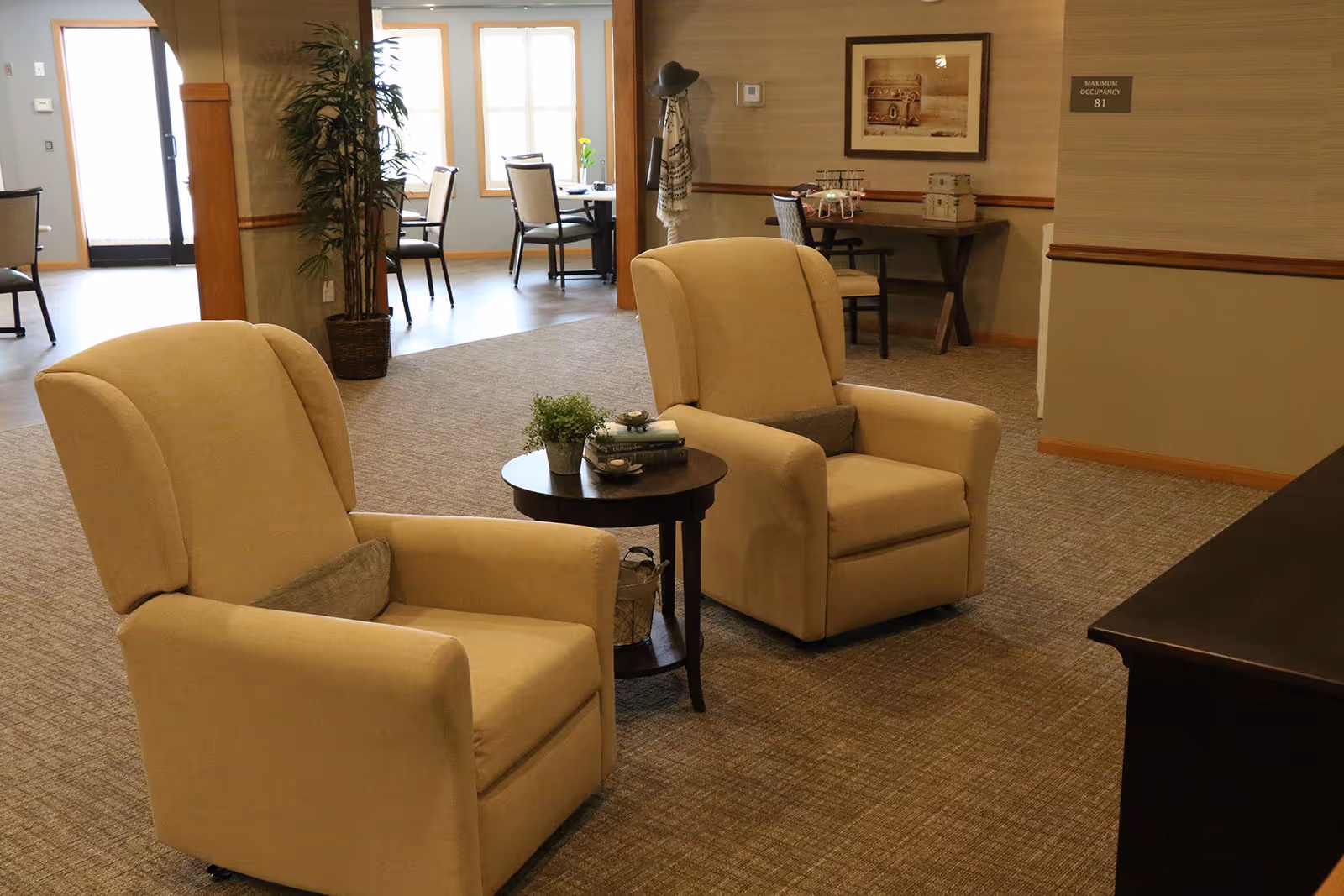 A cozy seating area in a senior living facility featuring two beige armchairs with small pillows, a round wooden side table with a small plant and books, and a carpeted floor. In the background, there are dining tables and chairs near large windows letting in natural light, a coat rack with a hat and scarf, and framed artwork on the wall.