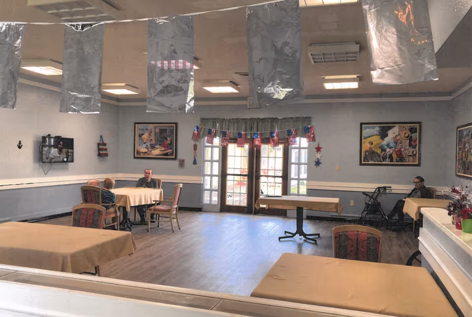 Spacious dining/activity room with tables and chairs, patriotic decorations, a wall-mounted TV, and a few seated residents.