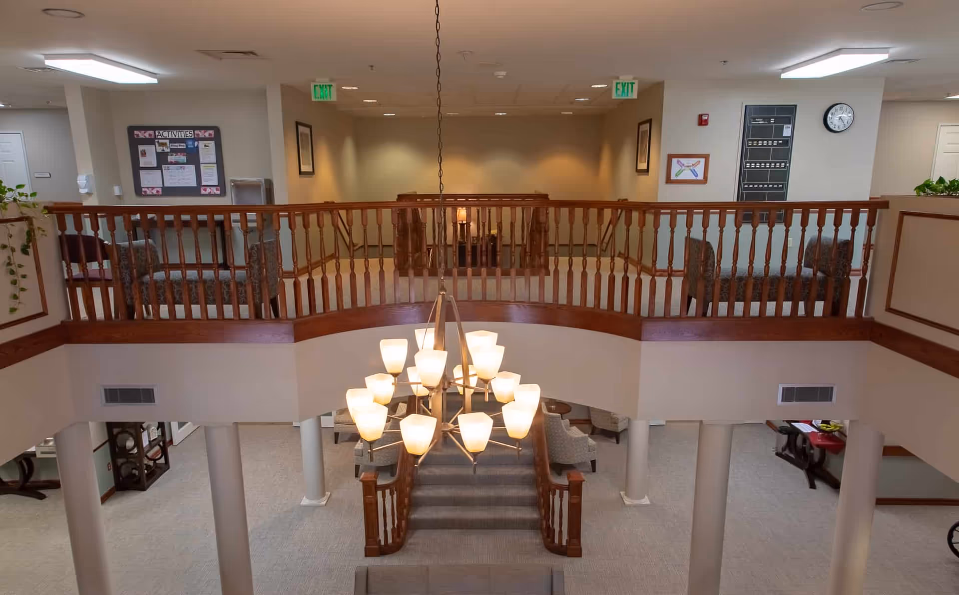 Interior view of a senior living facility showing a two-level common area with a wooden railing on the upper level, a large chandelier hanging in the center, and seating areas with chairs on both levels. The walls are light-colored, and there are exit signs and framed pictures visible.