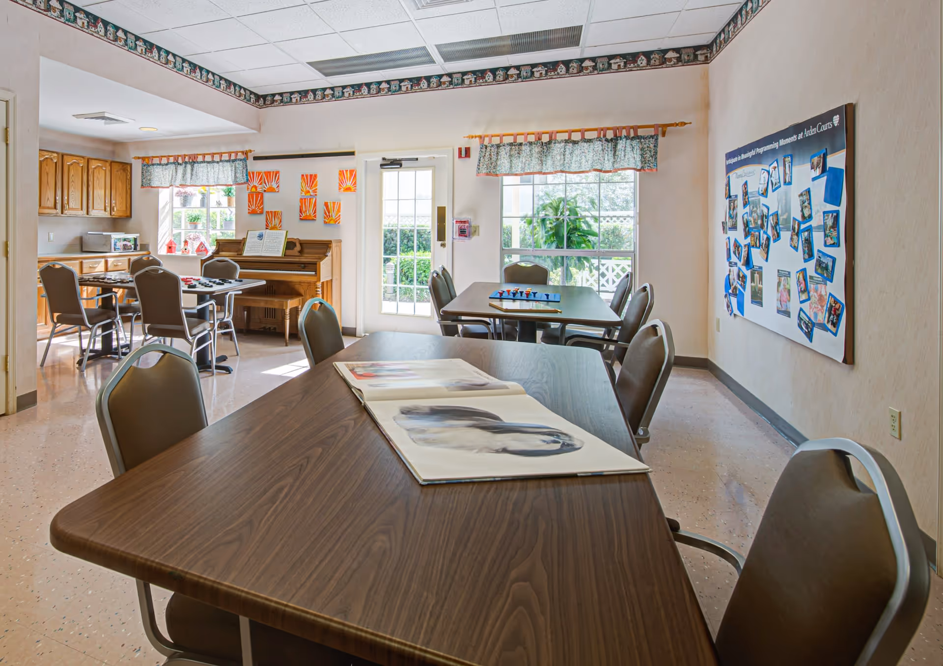 A bright room with several tables and chairs arranged for group activities. There is a piano against the wall with sheet music on it, colorful artwork on the walls, and a large bulletin board displaying photos and information. Windows with patterned valances let in natural light, and a door leads outside.