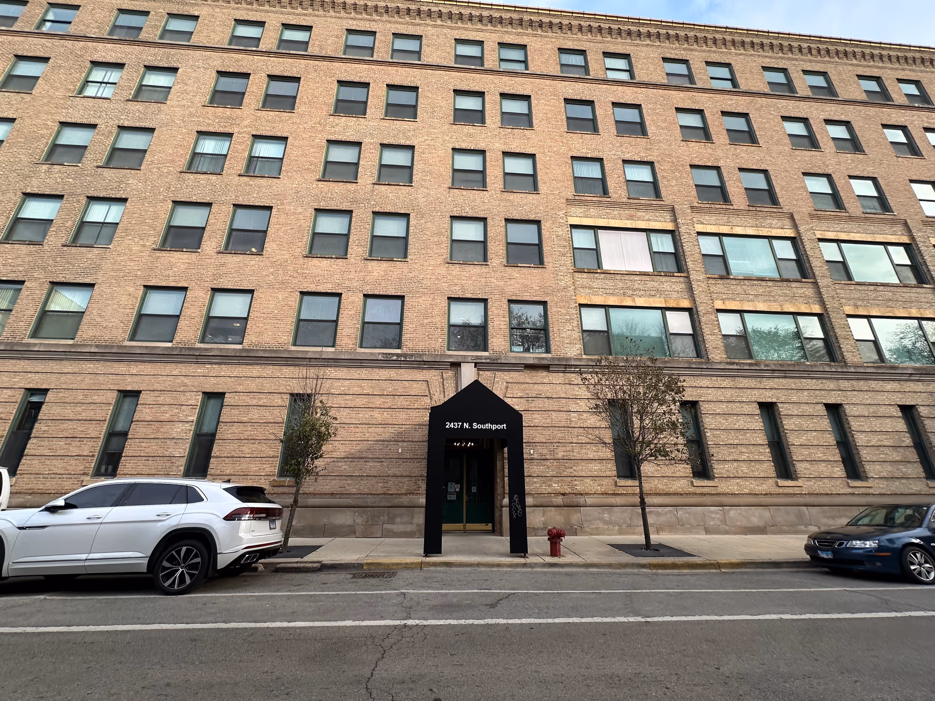 Exterior view of a multi-story brick building with many windows. There is a black awning over the entrance with the address '2437 N. Southport' displayed. Two cars are parked on the street in front of the building, along with two small trees and a red fire hydrant on the sidewalk.