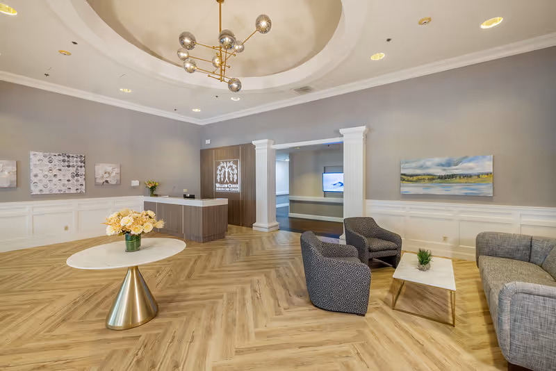 A spacious and well-lit reception area of Willow Creek Healthcare Center featuring a modern chandelier, a round table with a flower arrangement, a reception desk, two armchairs, a sofa, a coffee table with a small plant, and decorative paintings on the walls.