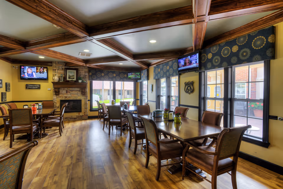 A cozy dining room with wooden floors, multiple tables and chairs, a stone fireplace, and several windows with patterned valances letting in natural light. Two flat-screen TVs are mounted on the walls, and the ceiling features wooden beams.