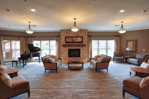 A spacious senior living lounge with a central fireplace, sofas, and a grand piano.
