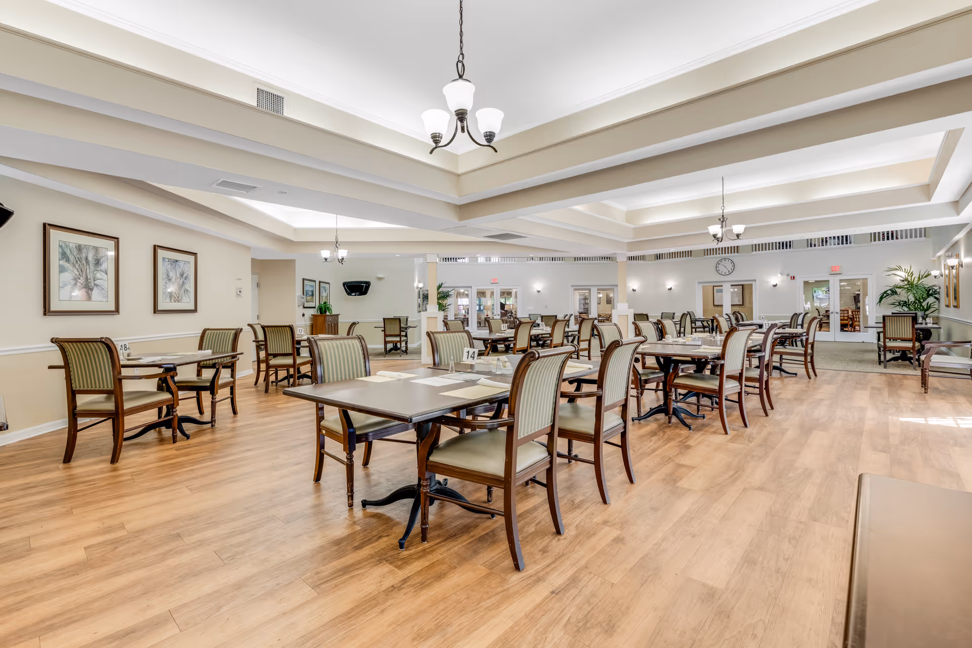 A spacious dining room with multiple wooden tables and chairs arranged neatly on a light wood floor. The room features a coffered ceiling with hanging light fixtures, framed artwork on the walls, and large windows allowing natural light. The area is clean and well-lit, designed for communal dining.