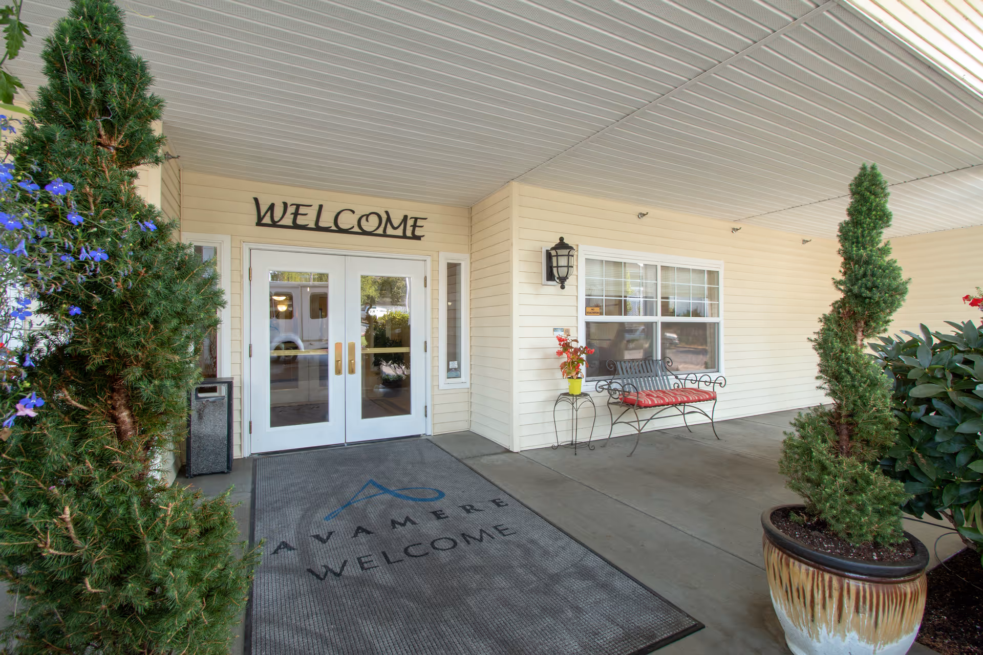 Entrance area of a senior living facility with double glass doors under a covered porch. Above the doors is a sign that says 'WELCOME'. There is a welcome mat on the ground with the text 'AVAMERE WELCOME'. To the right, there is a black metal bench with a red cushion and a small table with a yellow pot containing red flowers. Two large potted plants with green foliage frame the entrance.