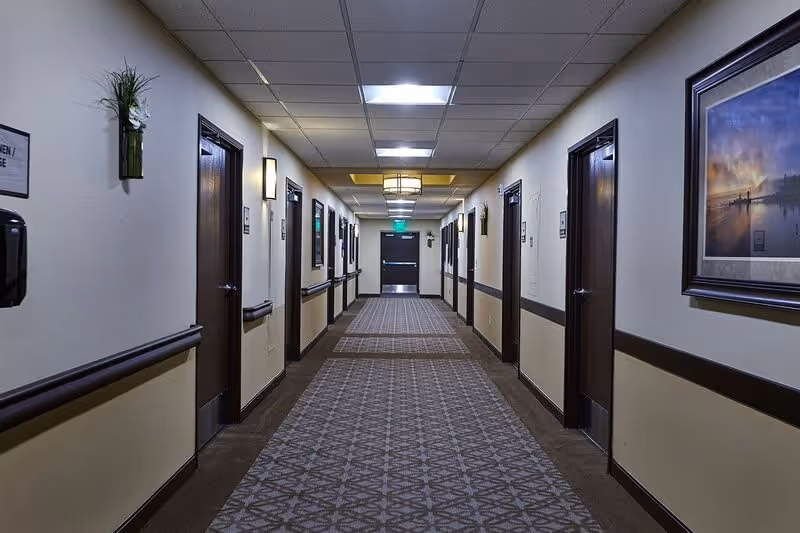 A long, well-lit hallway in an assisted living facility with multiple closed wooden doors on both sides. The walls are painted in a light color with a dark chair rail and decorated with framed artwork and wall-mounted plants. The ceiling has recessed lighting and a decorative light fixture in the center. At the end of the hallway, there is a double door with an exit sign above it.