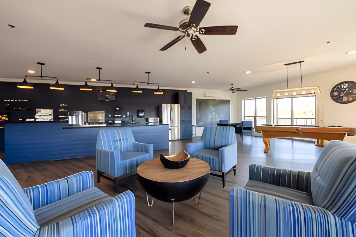 Open common lounge with striped armchairs arranged around a round coffee table, a bar with pendant lights, and a pool table by windows.