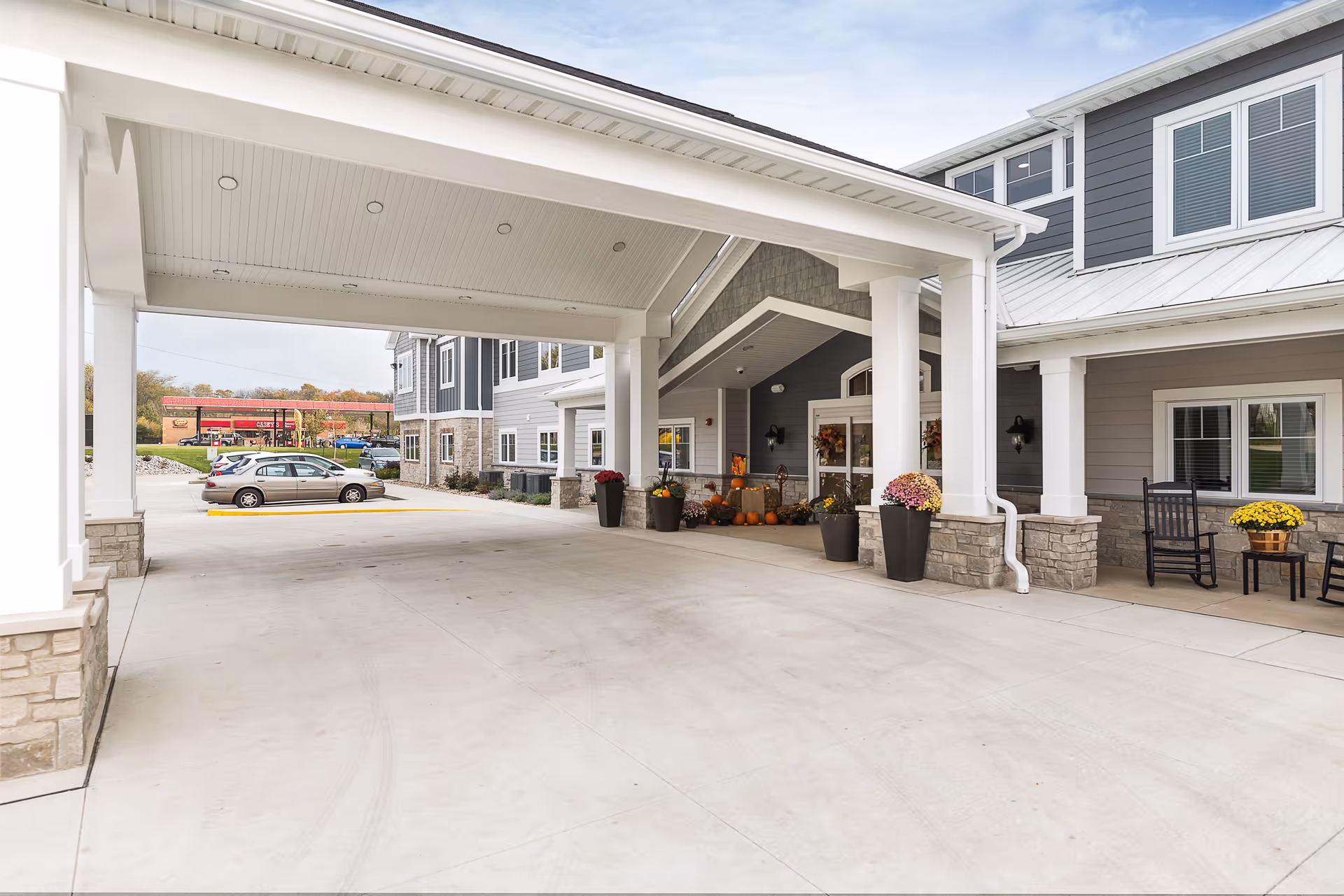Covered entrance area of Villas of Holly Brook Assisted Living & Memory Care building with stone and siding exterior, decorated with potted flowers and pumpkins. There are rocking chairs and a small table on the right side near windows. Cars are parked in the background near a gas station.