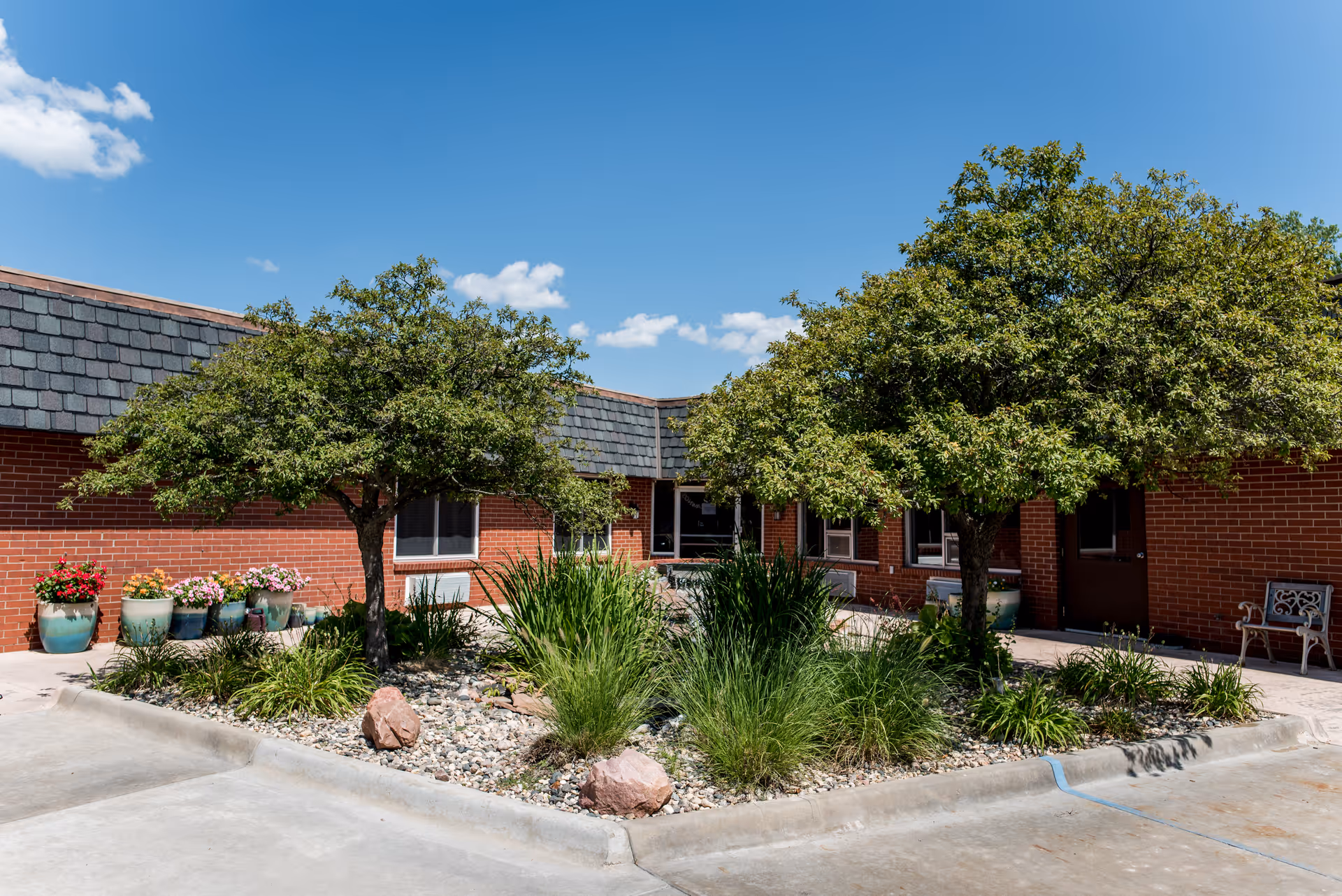 Courtyard in front of a single-story brick senior facility with two trees, planters, and landscaping under a clear blue sky.